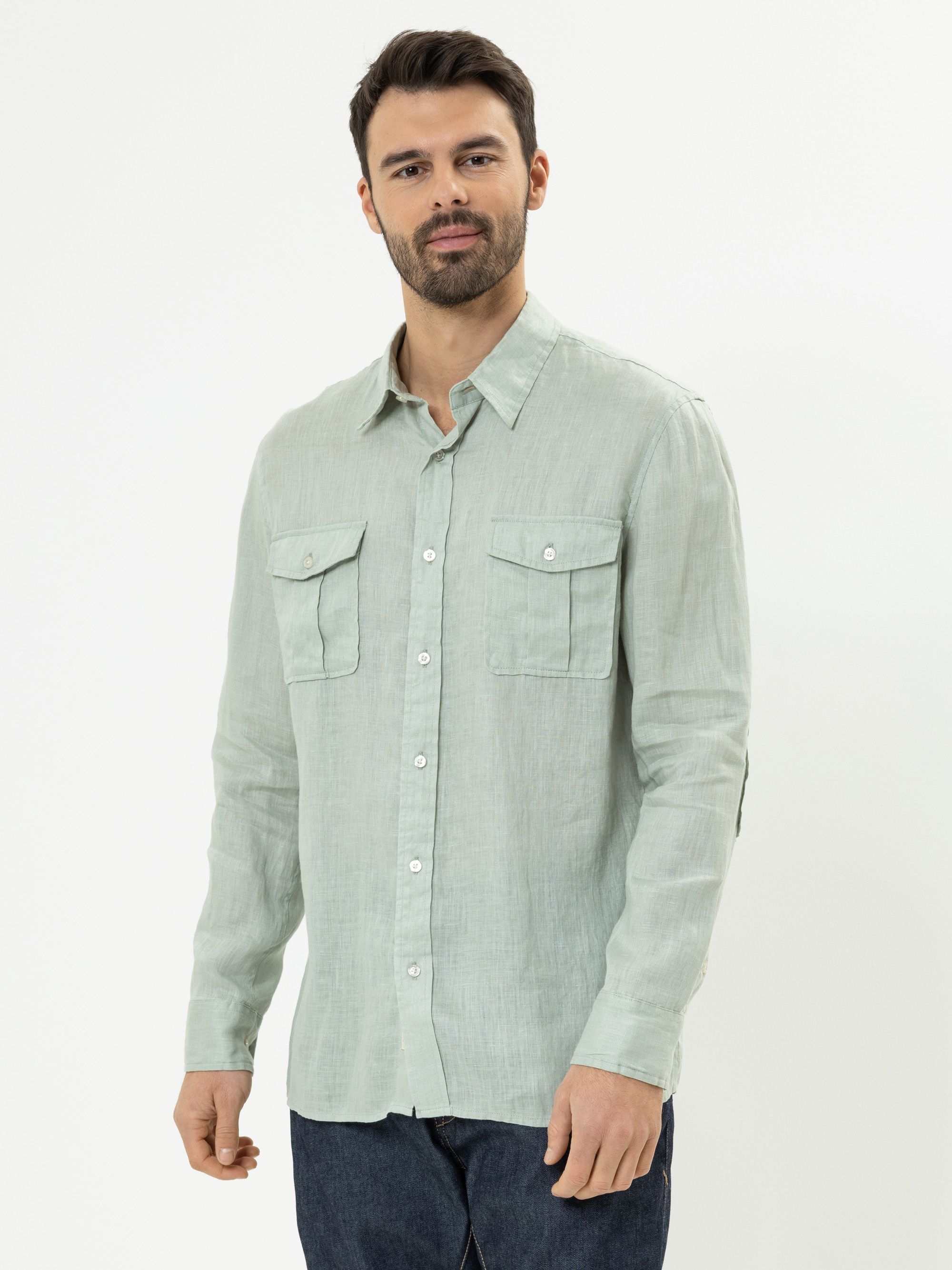 Ein Mann mit kurzen dunklen Haaren und Bart trägt ein hellgrünes Button-up-Hemd mit Brusttaschen und dunkle Jeans und steht vor einem schlichten weißen Hintergrund.