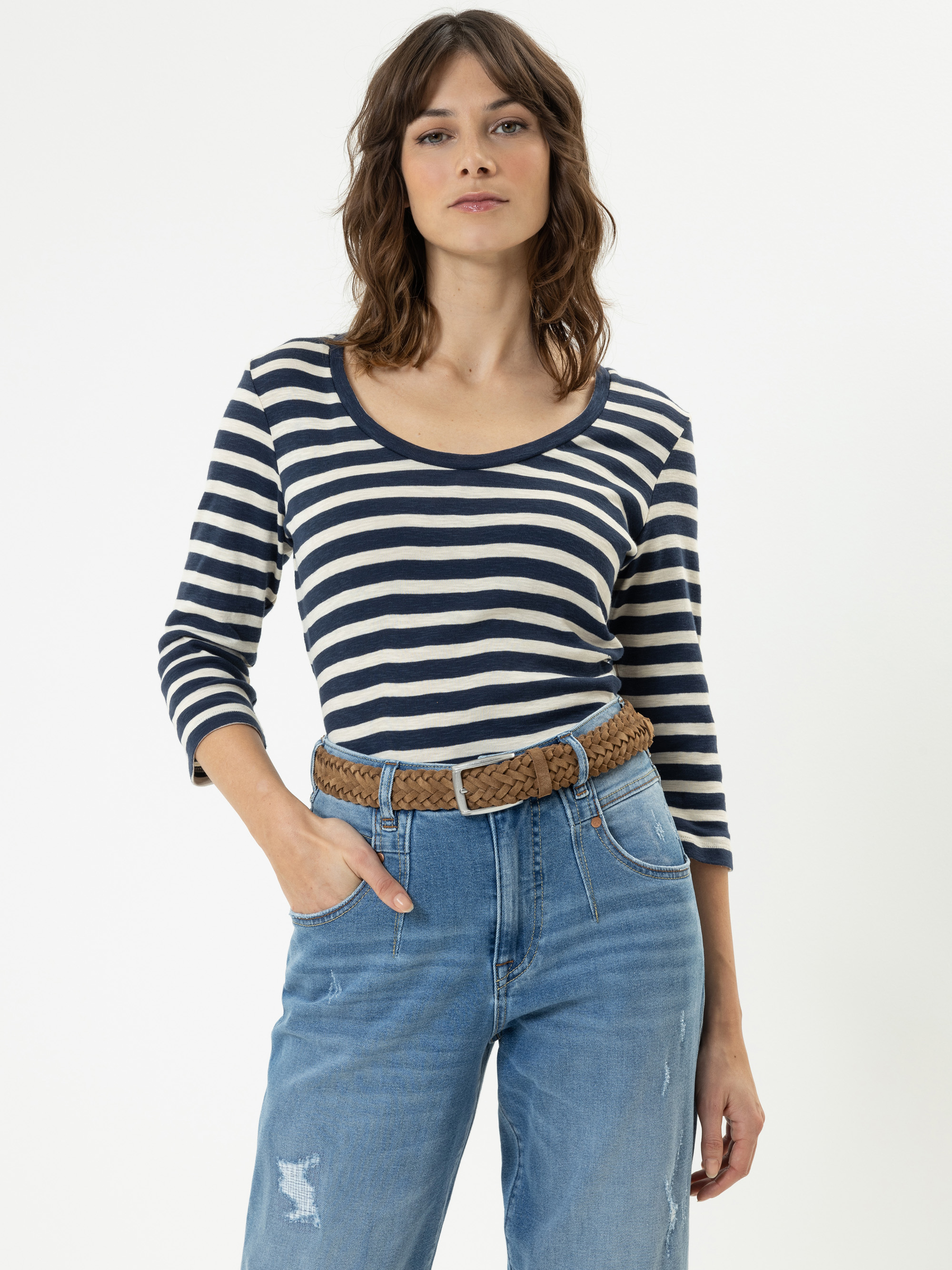 Frau mit gewelltem braunem Haar, die ein marineblau-weiß gestreiftes langärmeliges Hemd, blaue Jeans und einen geflochtenen braunen Gürtel trägt, steht vor einem schlichten weißen Hintergrund und hat eine Hand in der Tasche.