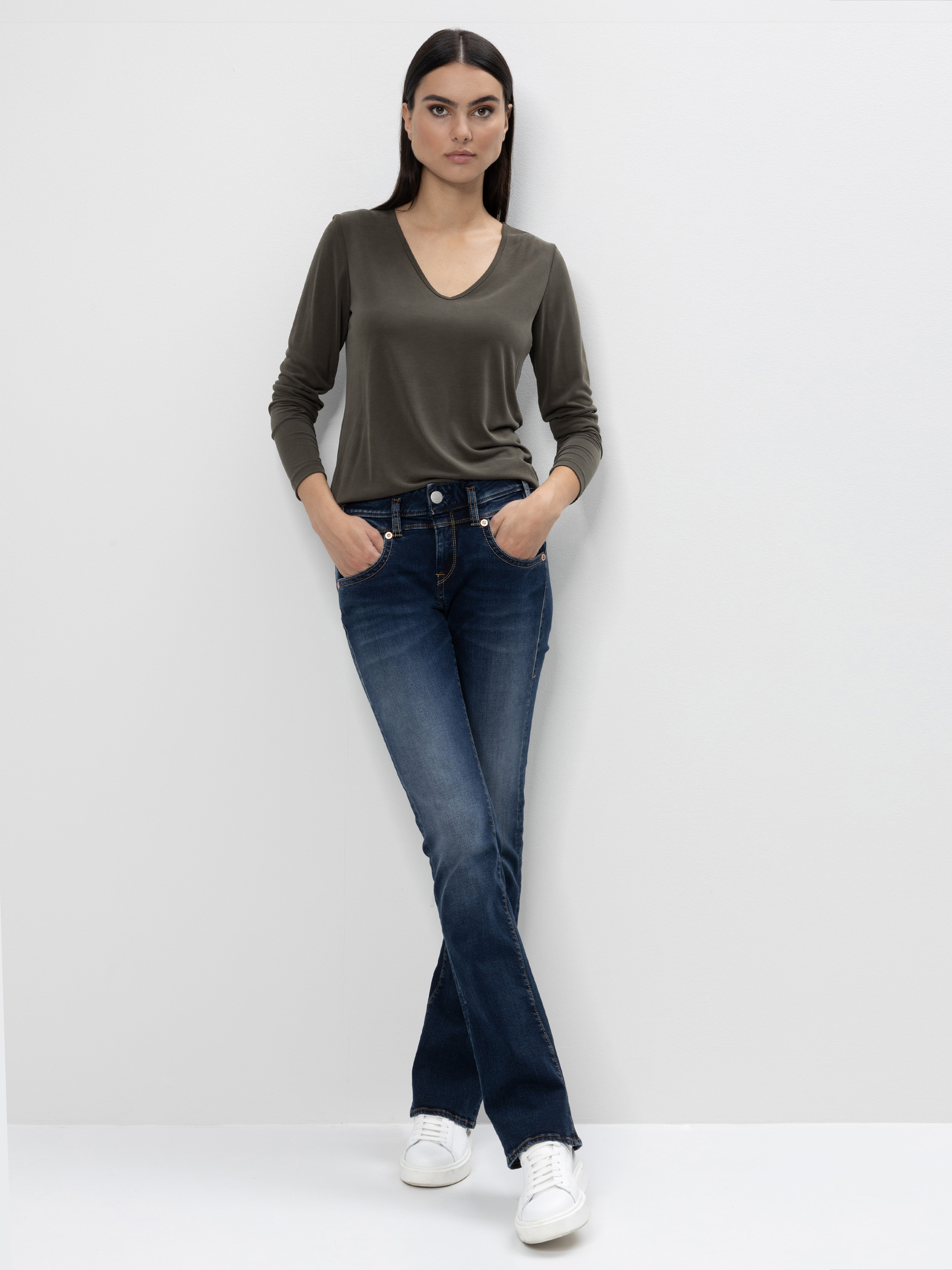 Eine Frau mit langen dunklen Haaren steht vor einem schlichten weißen Hintergrund. Sie trägt ein dunkelolivfarbenes langärmeliges Oberteil mit V-Ausschnitt, blaue Jeans und weiße Turnschuhe. Sie hat die Hände in den Taschen und schaut selbstbewusst i