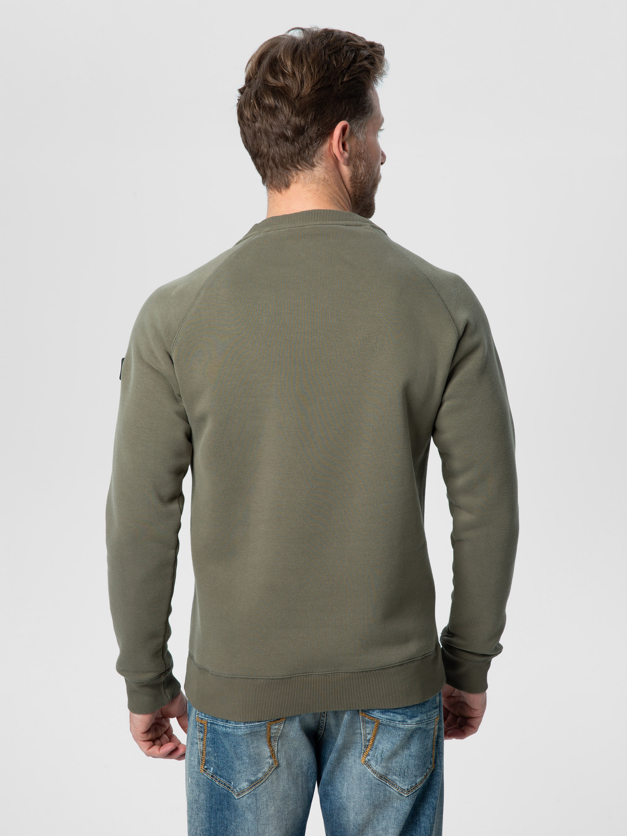 Ein Mann mit kurzen braunen Haaren, bekleidet mit einem olivgrünen Sweatshirt und blauen Jeans, steht mit dem Gesicht zur Kamera vor einem schlichten hellen Hintergrund.