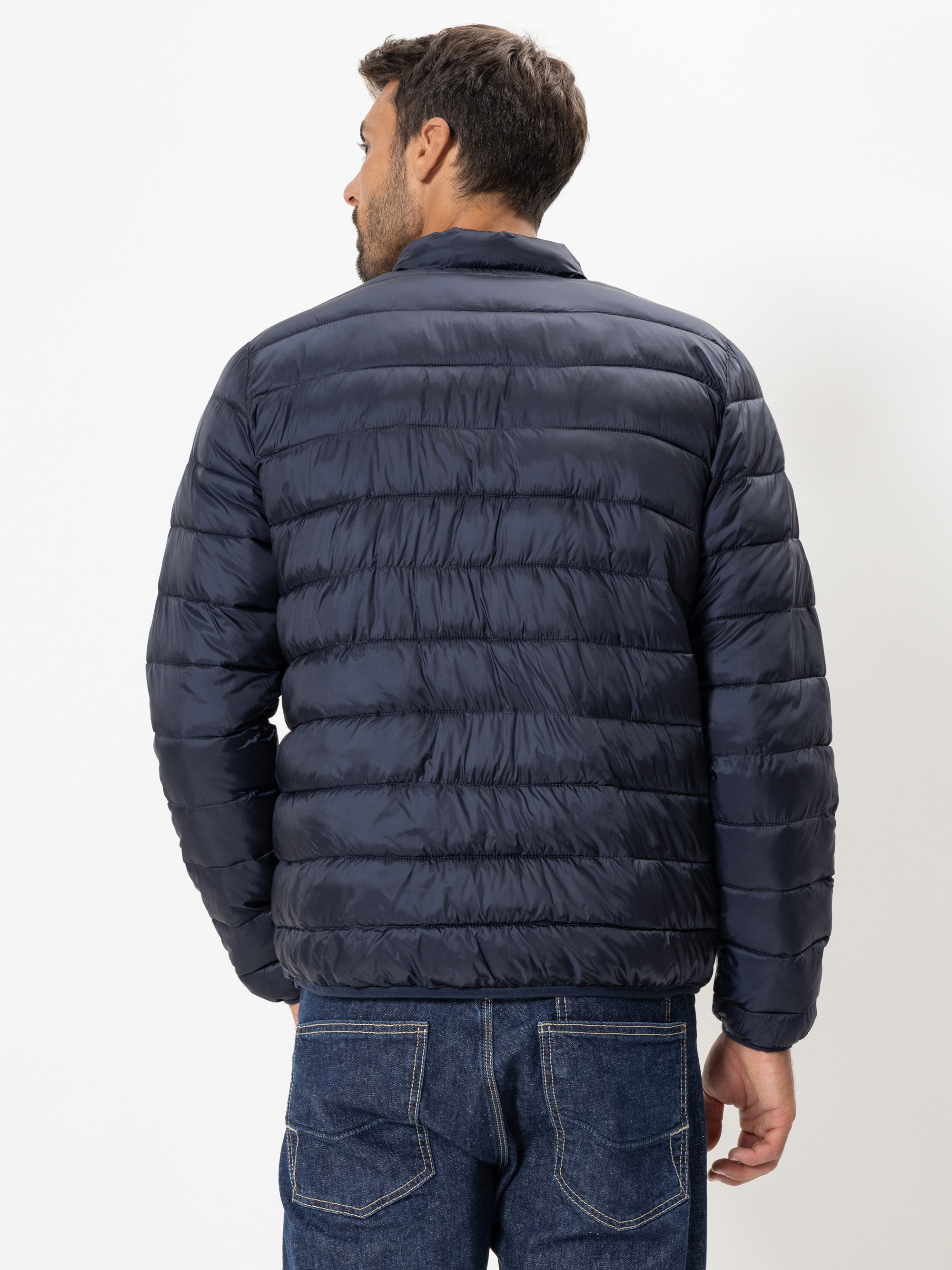 Ein Mann wird von hinten gezeigt, der eine marineblaue Pufferjacke und blaue Jeans trägt und vor einem schlichten weißen Hintergrund steht.