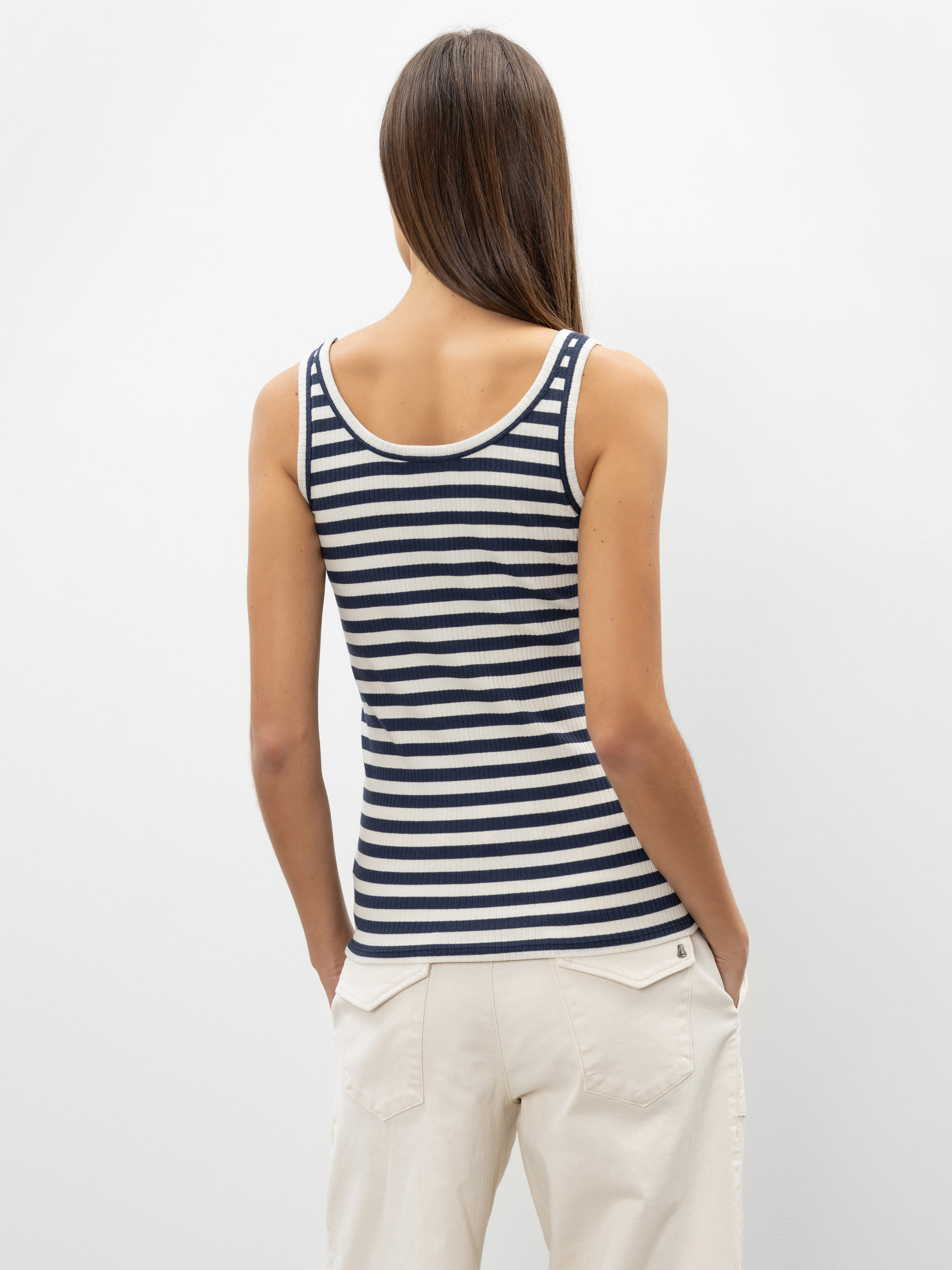 Eine Person mit langen braunen Haaren, von hinten gesehen, trägt ein marineblau-weiß gestreiftes ärmelloses Oberteil und eine cremefarbene Hose mit den Händen in den Taschen und steht vor einem schlichten hellen Hintergrund.