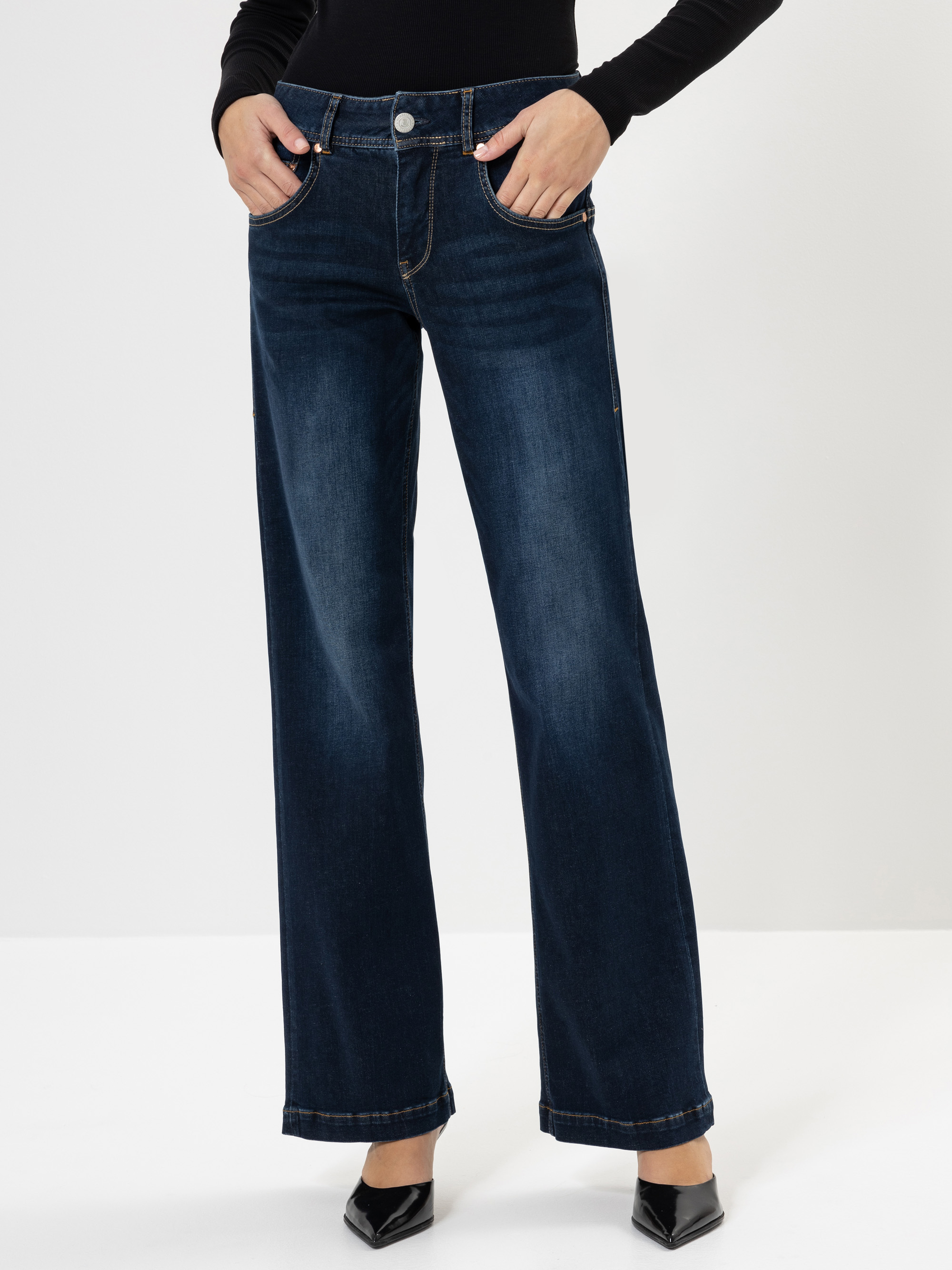Eine Person, die dunkelblaue Jeans mit weitem Bein und schwarze spitze Absätze trägt, steht vor einem schlichten weißen Hintergrund und hat die Hände in den Vordertaschen. Der Oberkörper ist nicht sichtbar.