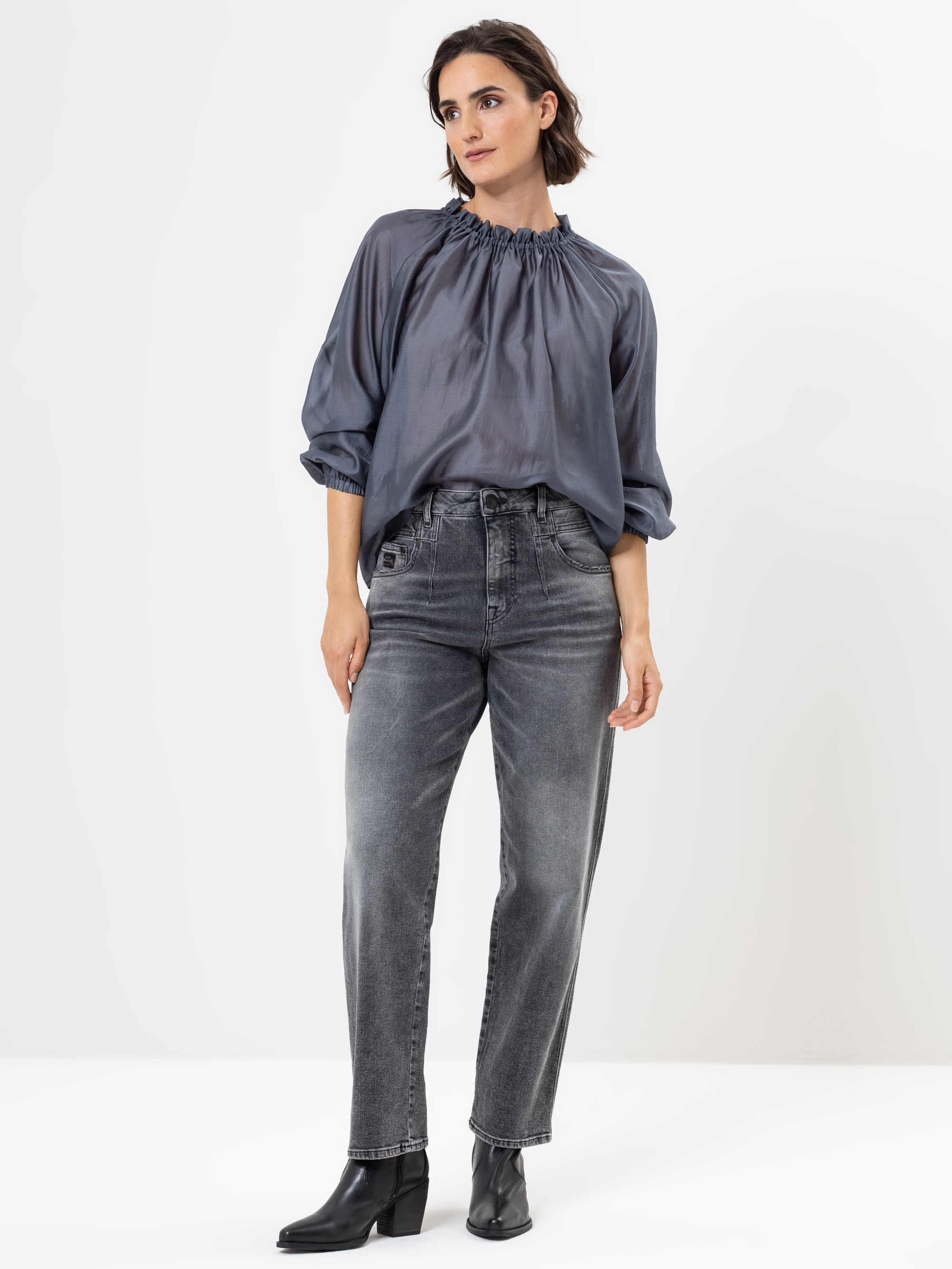 Eine Frau steht vor einem weißen Hintergrund und trägt eine lockere, glänzende graue Bluse mit gerafftem Ausschnitt, graue Jeans mit hoher Taille und schwarze Ankle Boots. Sie hat kurzes dunkles Haar und eine entspannte, selbstbewusste Pose.