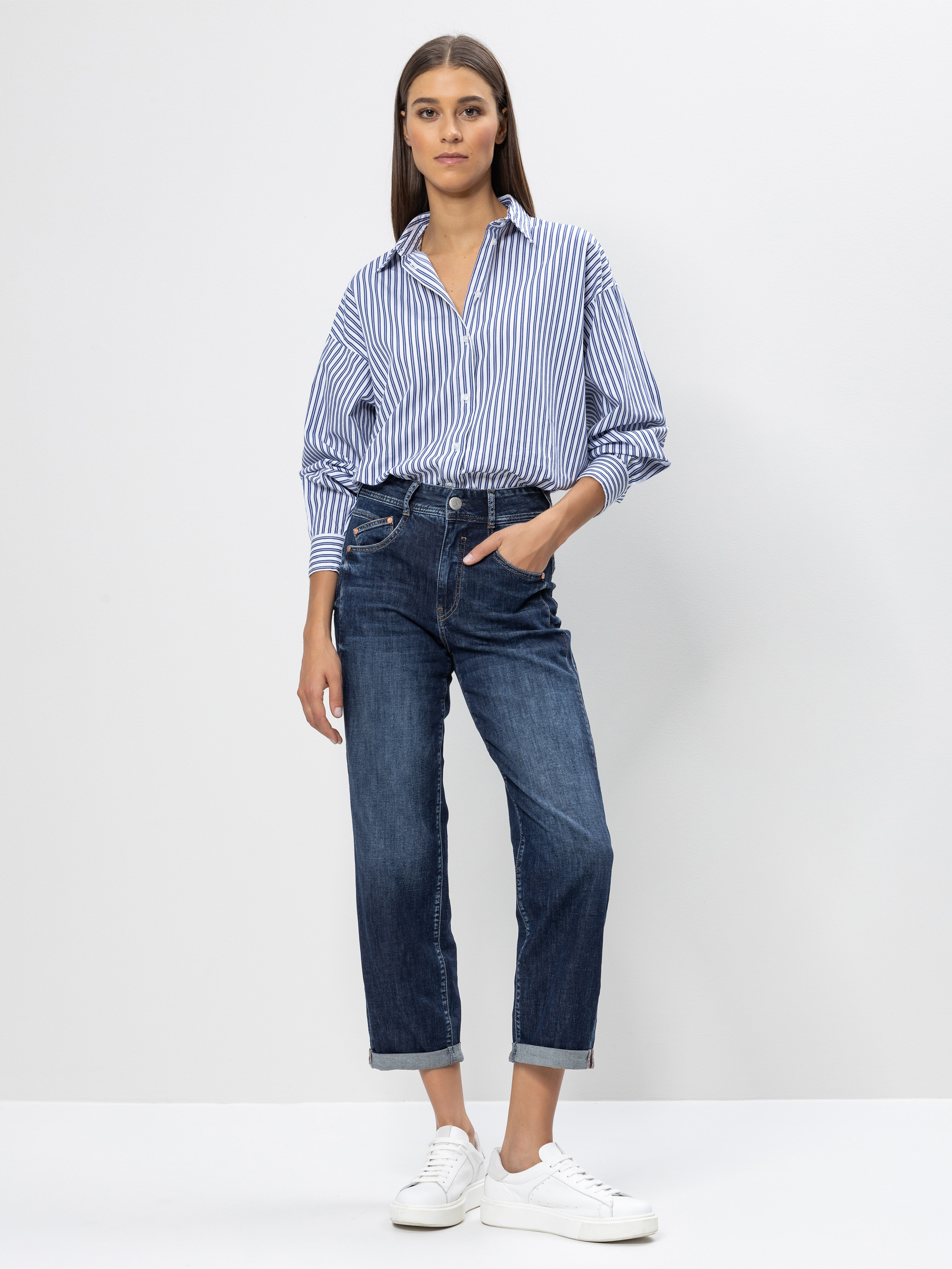 Eine Frau steht vor einem schlichten weißen Hintergrund, trägt ein blau-weiß gestreiftes Button-up-Hemd, dunkle Jeans mit Manschetten und weiße Turnschuhe, eine Hand in der Tasche.