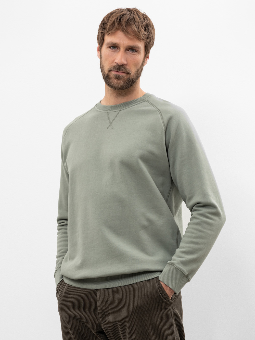 Ein Mann mit braunem Haar und Bart trägt ein hellgrünes Sweatshirt und eine braune Cordhose und steht mit den Händen in den Taschen vor einem schlichten weißen Hintergrund.
