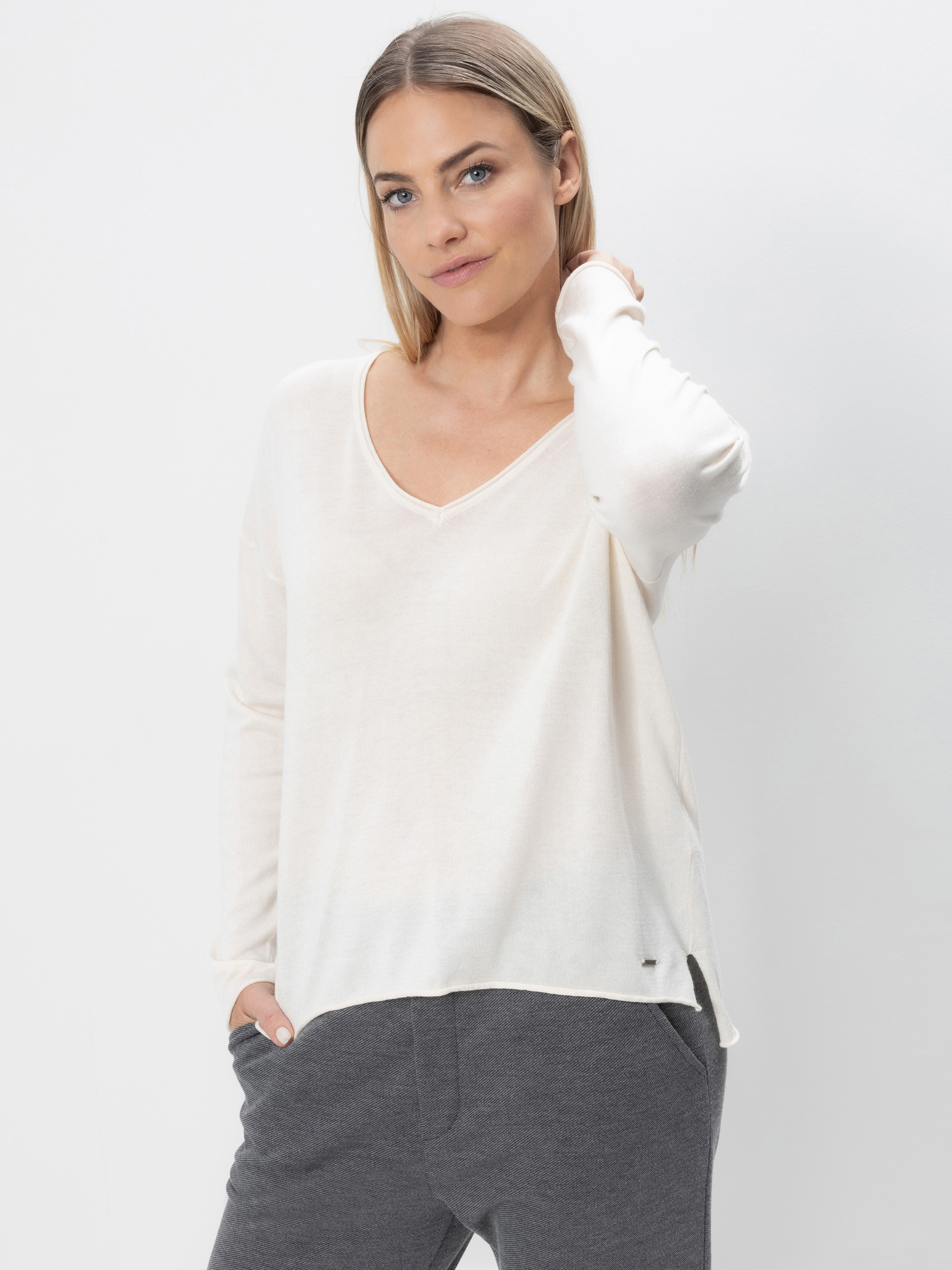 Eine Frau mit glattem, hellbraunem Haar trägt ein lockeres, langärmeliges weißes Oberteil und eine dunkelgraue Hose. Sie steht vor einem schlichten weißen Hintergrund und hat eine Hand in der Tasche, während die andere ihren Hals berührt.
