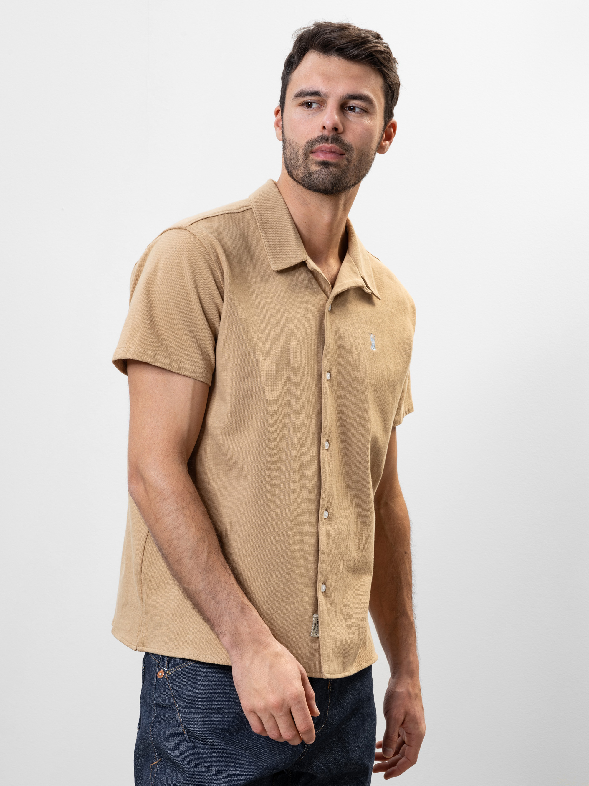 Ein Mann mit kurzen dunklen Haaren, der ein beigefarbenes kurzärmeliges Button-up-Hemd und dunkle Jeans trägt, steht vor einem schlichten weißen Hintergrund und blickt leicht nach links.