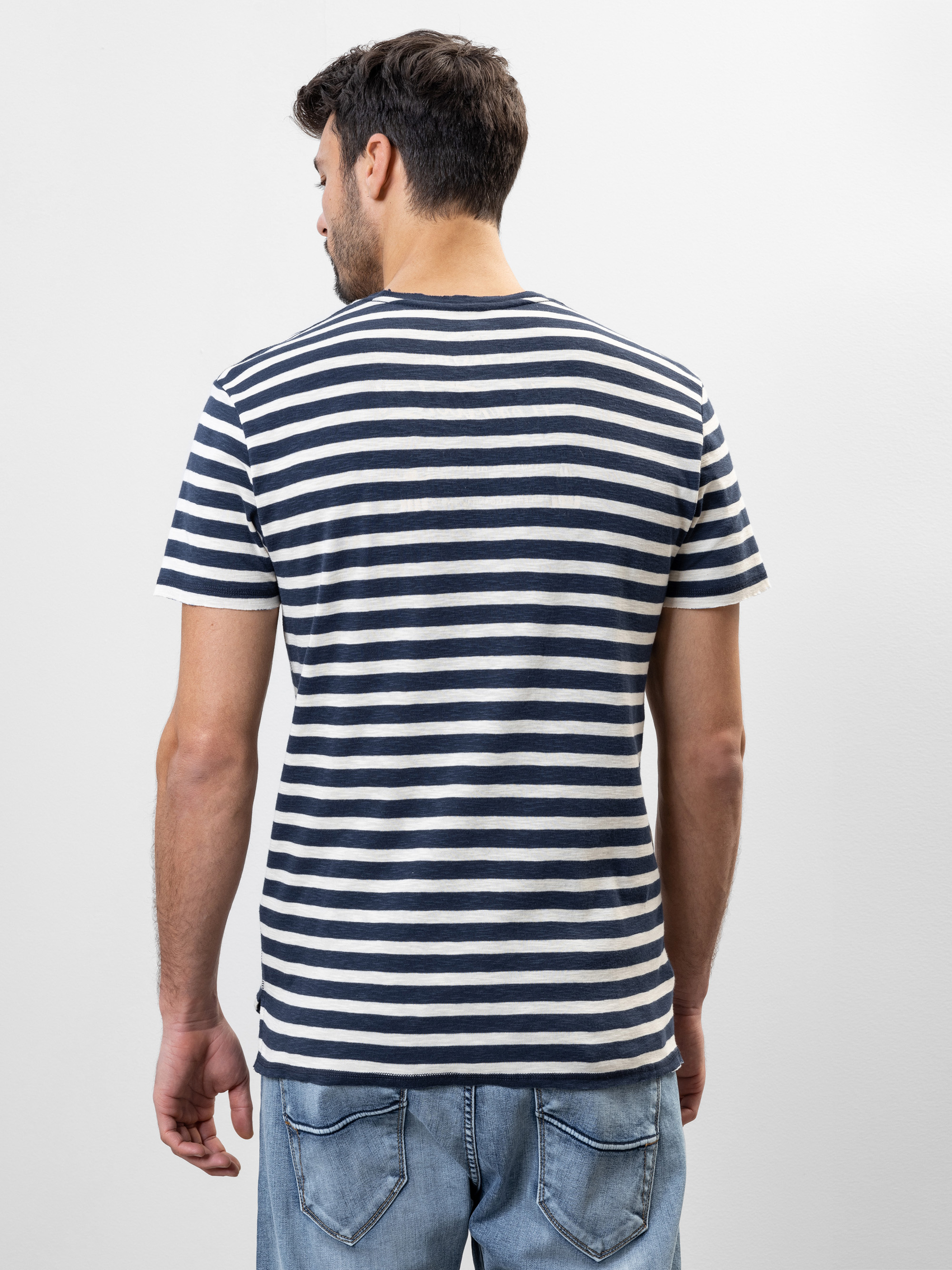 Ein Mann, der ein marineblau-weiß gestreiftes T-Shirt und hellblaue Jeans trägt, steht mit dem Gesicht von der Kamera abgewandt vor einem schlichten weißen Hintergrund.