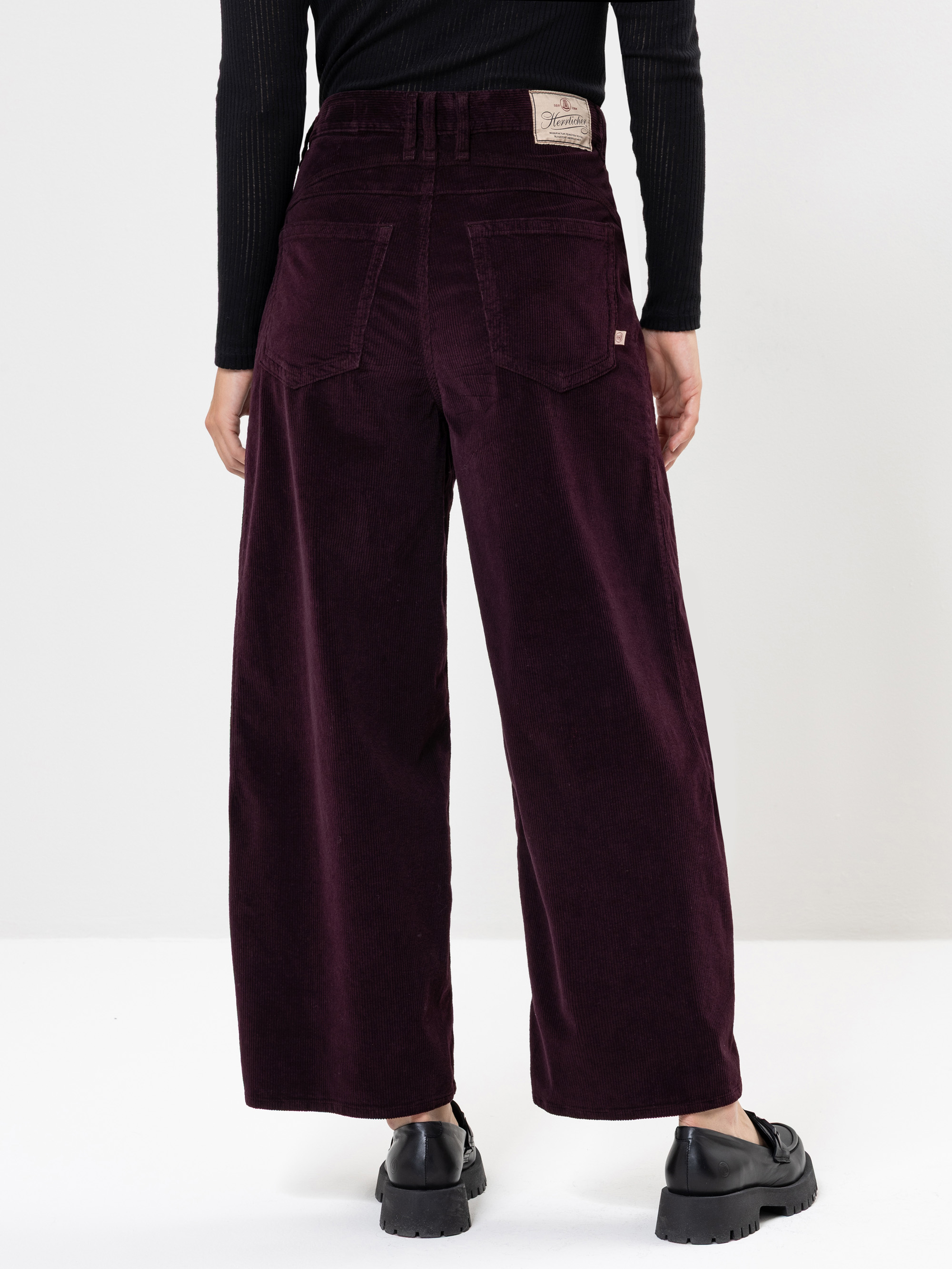 Eine Person trägt eine dunkellila Cordhose mit weitem Bein und schwarze Schuhe, von hinten gesehen. Die Hose hat Gesäßtaschen, einen Lederaufnäher am Bund und wird mit einem schwarzen, langärmeligen Oberteil kombiniert.