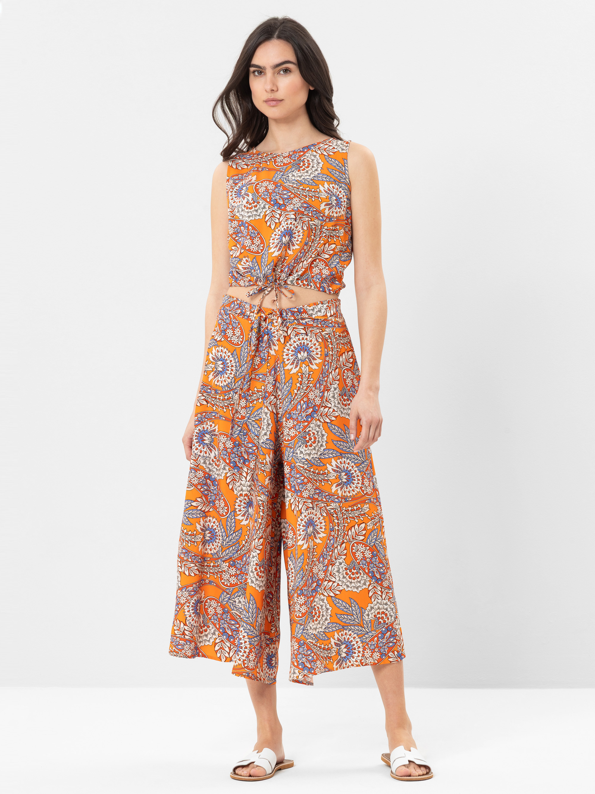Eine Frau steht vor einem unifarbenen Hintergrund und trägt ein ärmelloses, orangefarbenes und mehrfarbig gemustertes Crop-Top und eine dazu passende Hose mit weitem Bein, dazu weiße Sandalen.