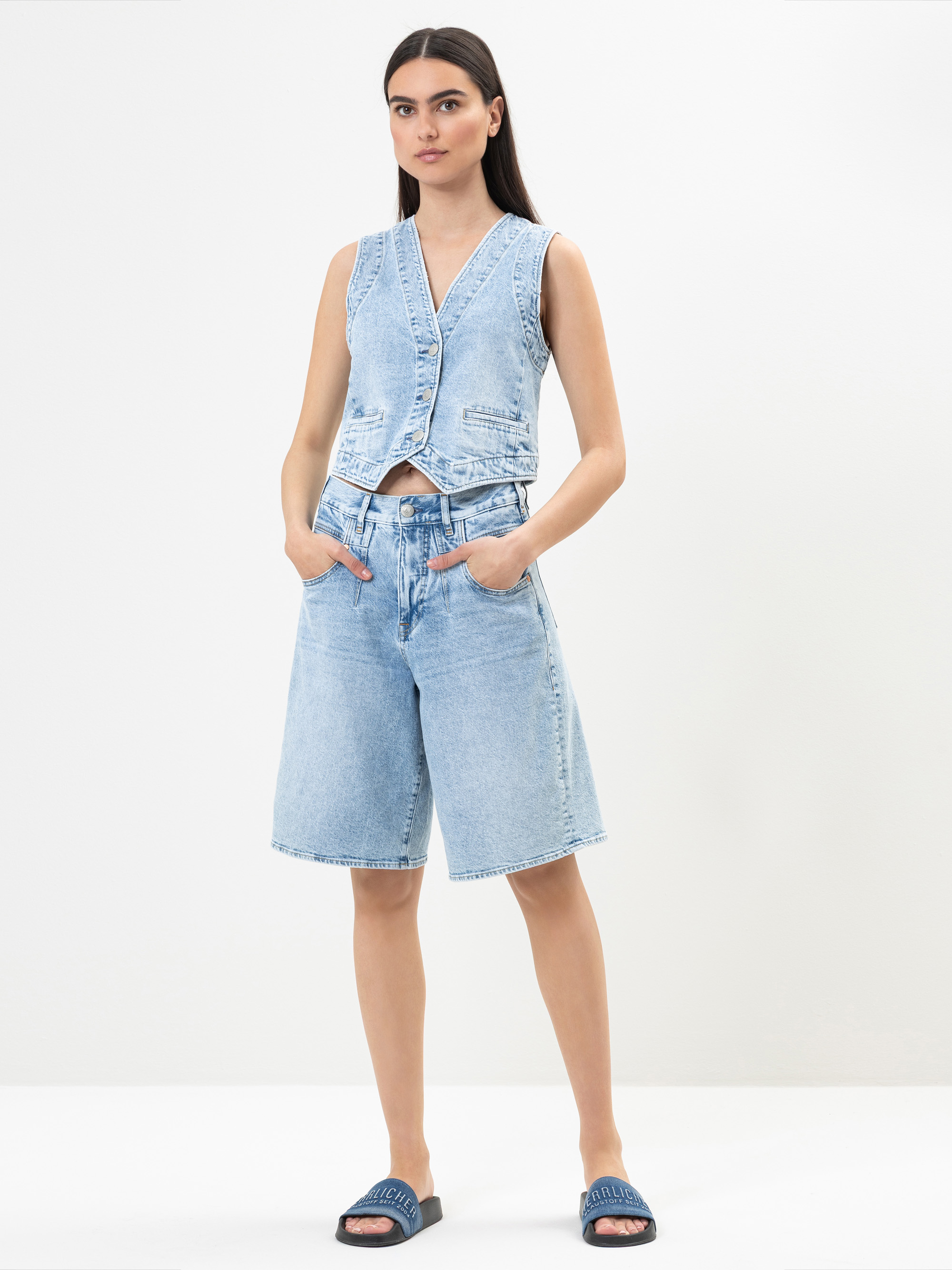 Eine Frau steht vor einem weißen Hintergrund und trägt eine ärmellose hellblaue Jeansweste, dazu passende knielange Jeansshorts und blaue Slide-Sandalen, wobei sie die Hände in den Taschen hat.