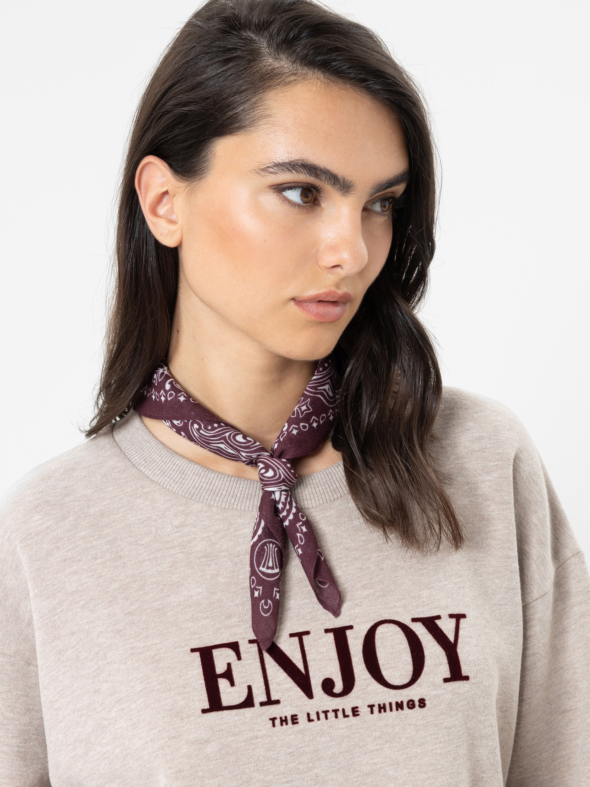 Eine Frau mit dunklem Haar, die ein beiges Sweatshirt mit der Aufschrift „ENJOY THE LITTLE THINGS“ in dunkelroter Schrift trägt und ein lila Bandana um den Hals gebunden hat; sie blickt zur Seite vor einem schlichten Hintergrund.