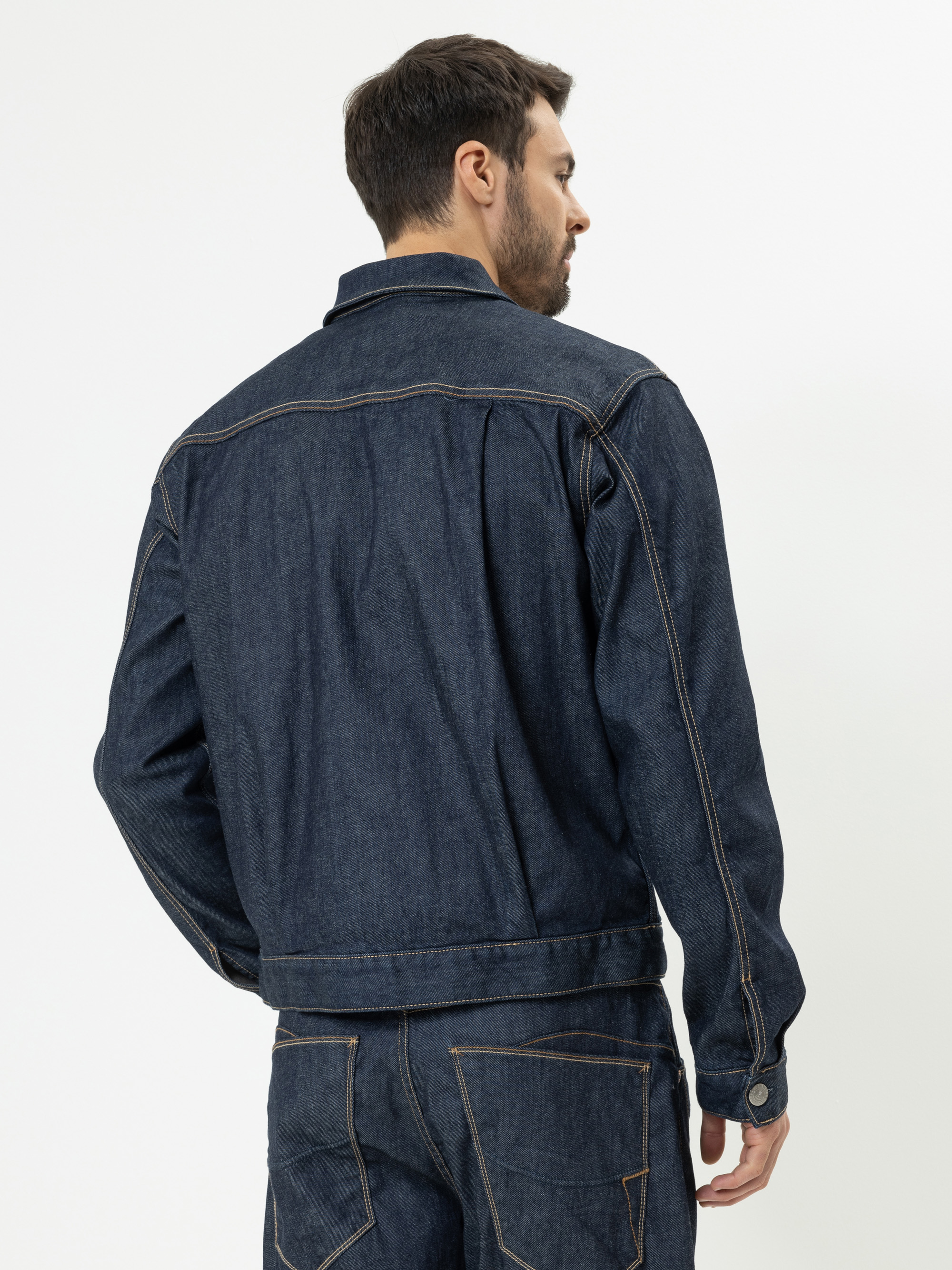 Ein Mann mit kurzen Haaren, von hinten gesehen, trägt eine dunkelblaue Jeansjacke und passende Jeans. Er steht vor einem schlichten weißen Hintergrund.