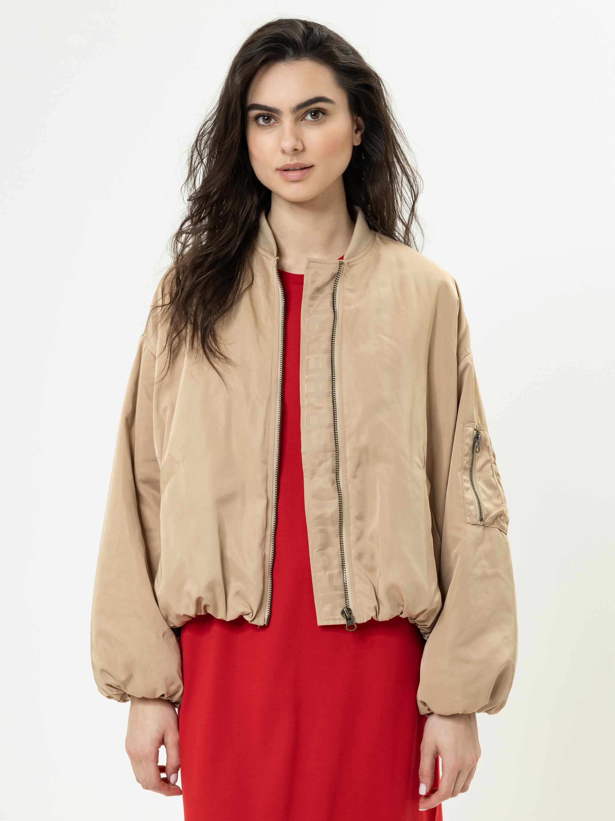 Eine Frau mit langen dunklen Haaren trägt eine hellbraune Bomberjacke über einem roten Kleid, steht vor einem schlichten weißen Hintergrund und blickt mit neutralem Blick in die Kamera.
