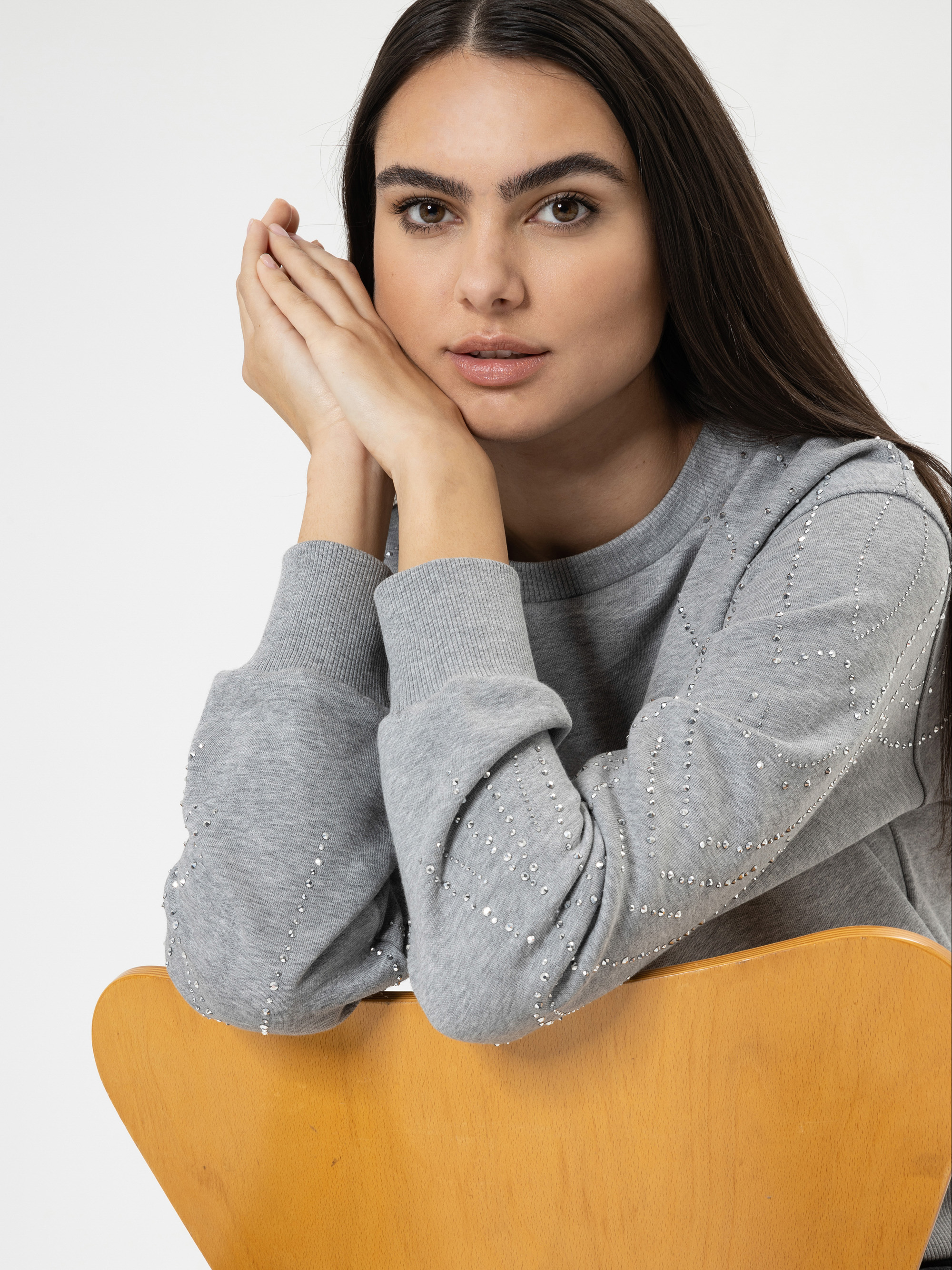 Eine Frau mit langen dunklen Haaren sitzt mit dem Rücken zugewandt auf einem Holzstuhl und stützt ihre Arme und ihr Kinn auf die Stuhllehne. Sie trägt einen hellgrauen Pullover mit dekorativen Verzierungen und blickt ruhig in die Kamera.