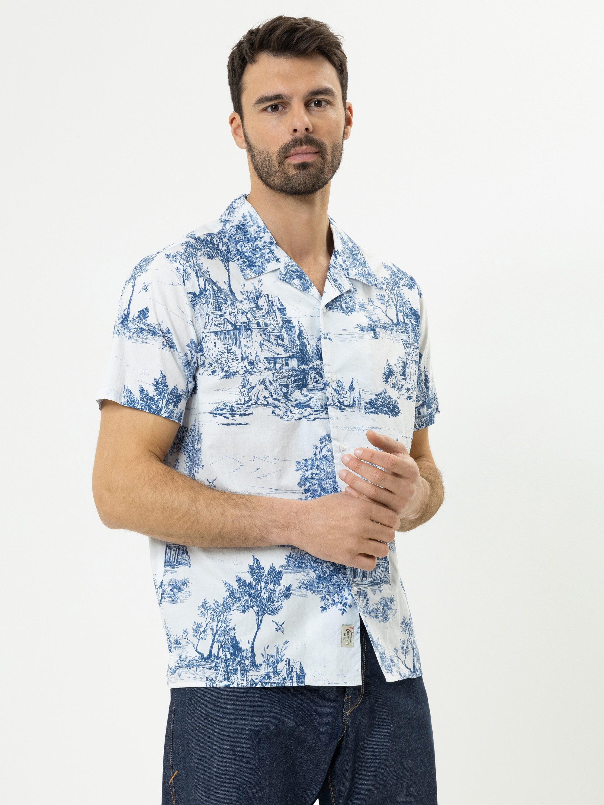 Ein Mann mit kurzen dunklen Haaren und einem Bart trägt ein weißes kurzärmeliges Hemd mit blauem Hirtenaufdruck und dunkle Jeans vor einem schlichten weißen Hintergrund.