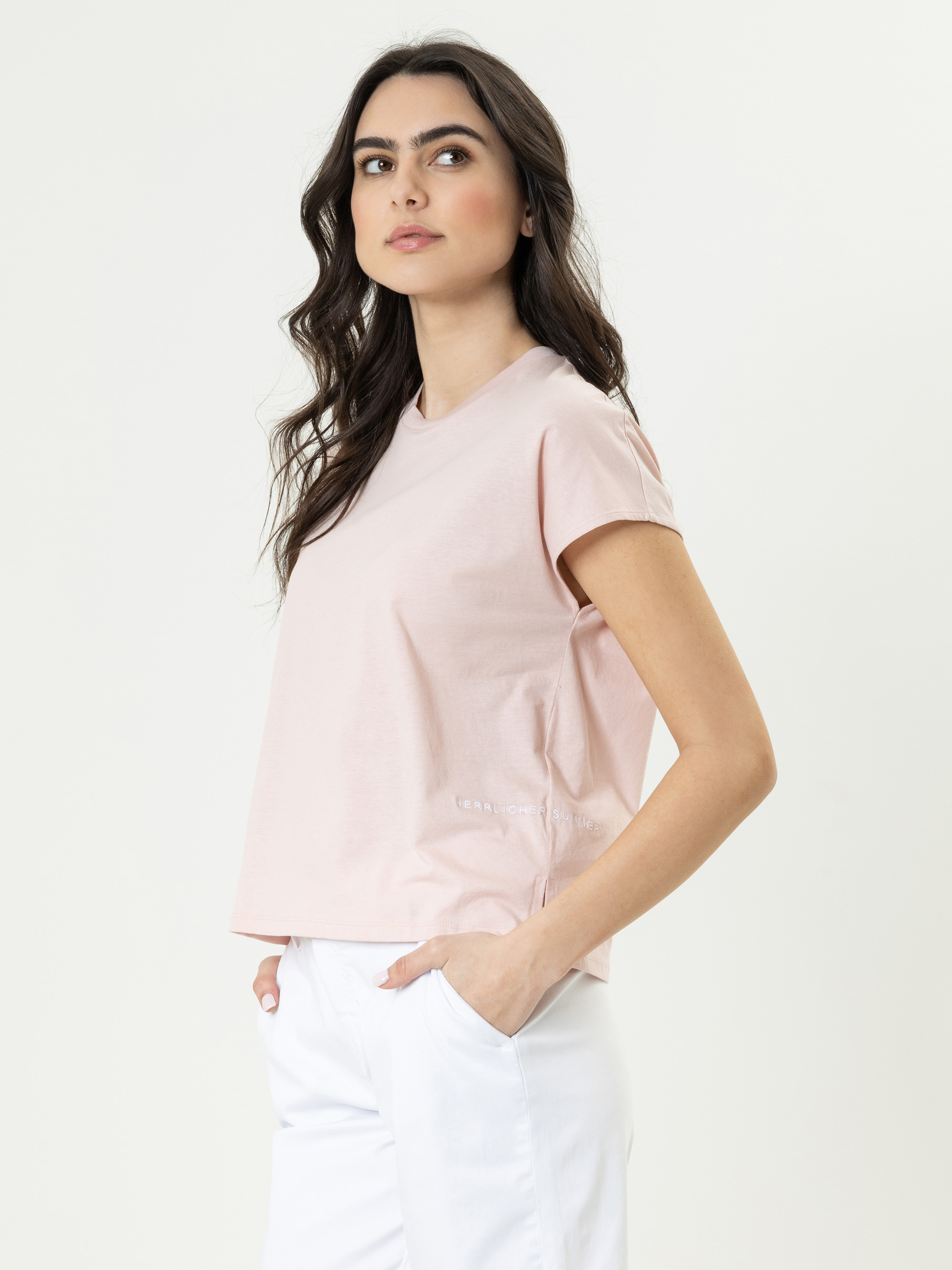 Eine Frau mit langen dunklen Haaren, die ein hellrosa T-Shirt und eine weiße Hose trägt, steht seitlich mit den Händen in den Taschen und blickt leicht nach oben vor einem schlichten hellen Hintergrund.