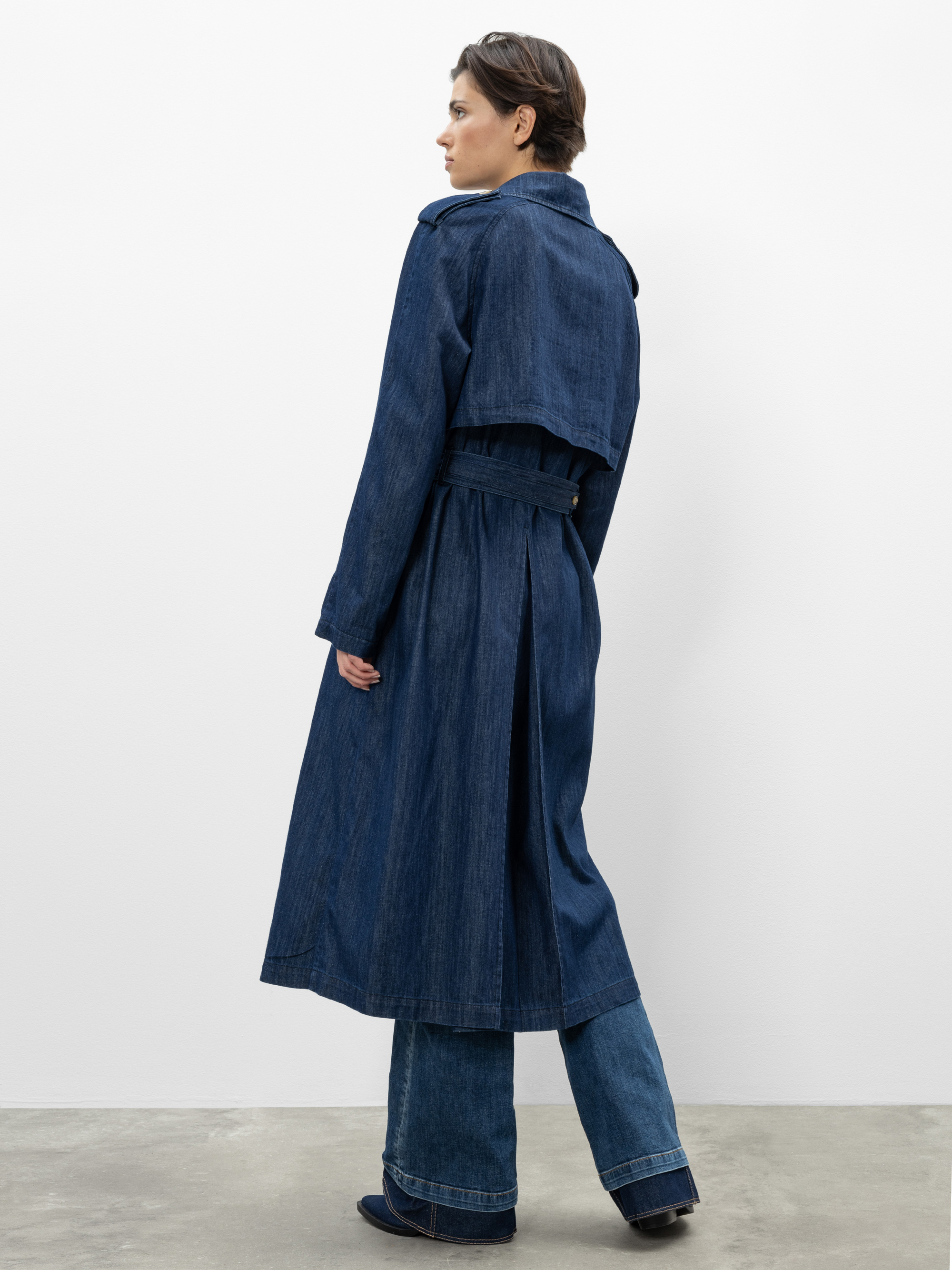 Eine Person mit kurzen Haaren, von hinten gesehen, trägt einen langen, gegürteten Jeans-Trenchcoat über Schlagjeans und dunkle Schuhe und steht auf einem grauen Boden vor einer schlichten weißen Wand.