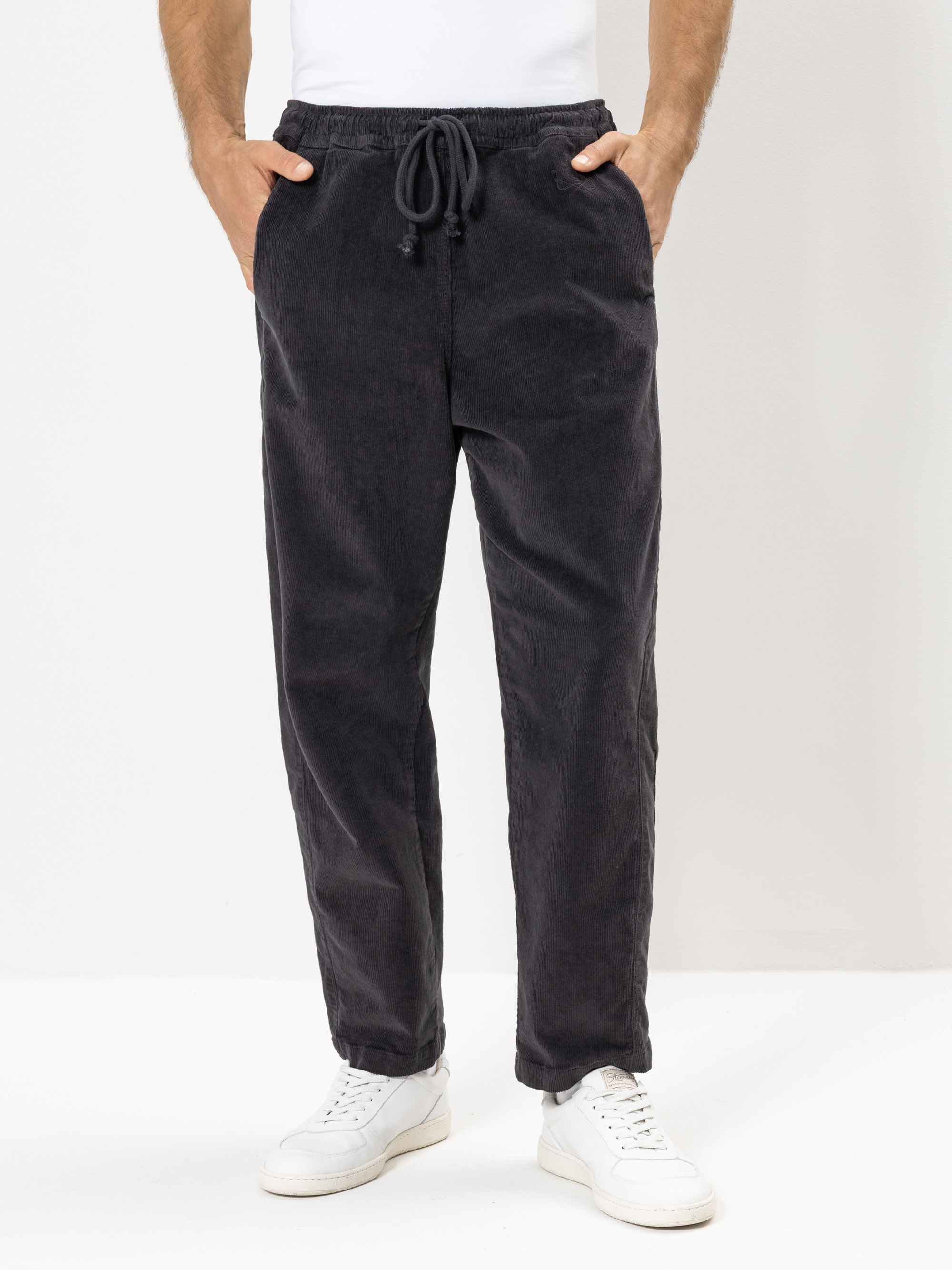 Eine Person, die eine locker sitzende, dunkelgraue Hose mit Kordelzug und weiße Turnschuhe trägt, steht vor einem schlichten weißen Hintergrund. Ihre Hände stecken in den Taschen, und nur ihr Unterkörper ist zu sehen.