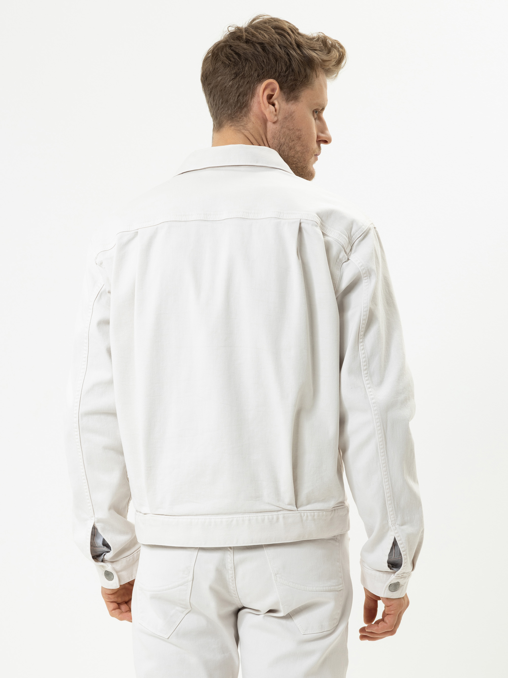 Ein Mann mit kurzen, hellbraunen Haaren steht mit dem Rücken zur Kamera, trägt eine weiße Jeansjacke und dazu passende weiße Jeans vor einem schlichten weißen Hintergrund.