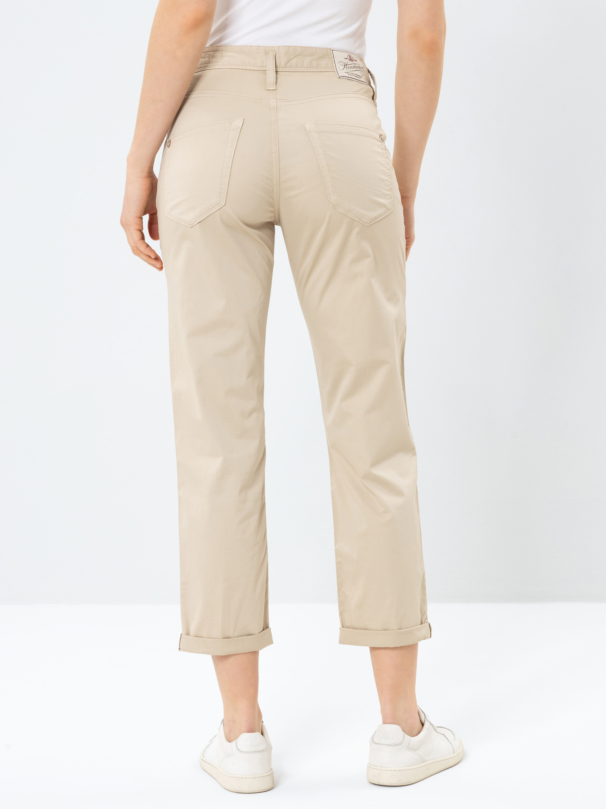 Eine Person, die eine hellbeige Hose mit geradem Bein und aufgerollten Manschetten sowie weiße Turnschuhe trägt, wird von hinten vor einem schlichten weißen Hintergrund gezeigt. Die Hose hat Gesäßtaschen und wird mit einem weißen Oberteil kombiniert