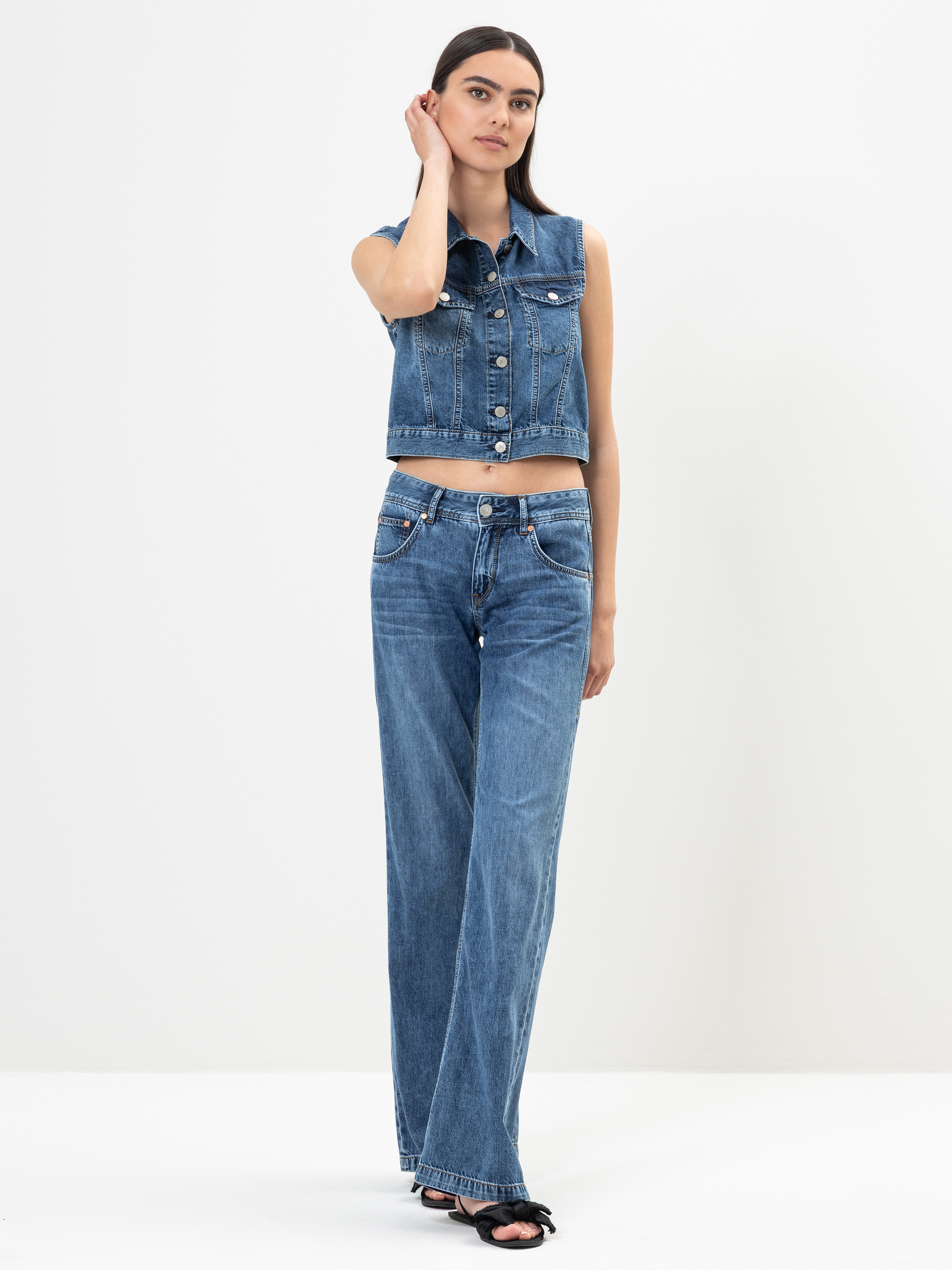 Eine Frau steht vor einem schlichten weißen Hintergrund und trägt eine ärmellose Jeansweste, eine hochgeschnittene Jeans mit weitem Bein und schwarze Sandalen. Sie hat glattes dunkles Haar und stützt eine Hand seitlich auf ihren Kopf.