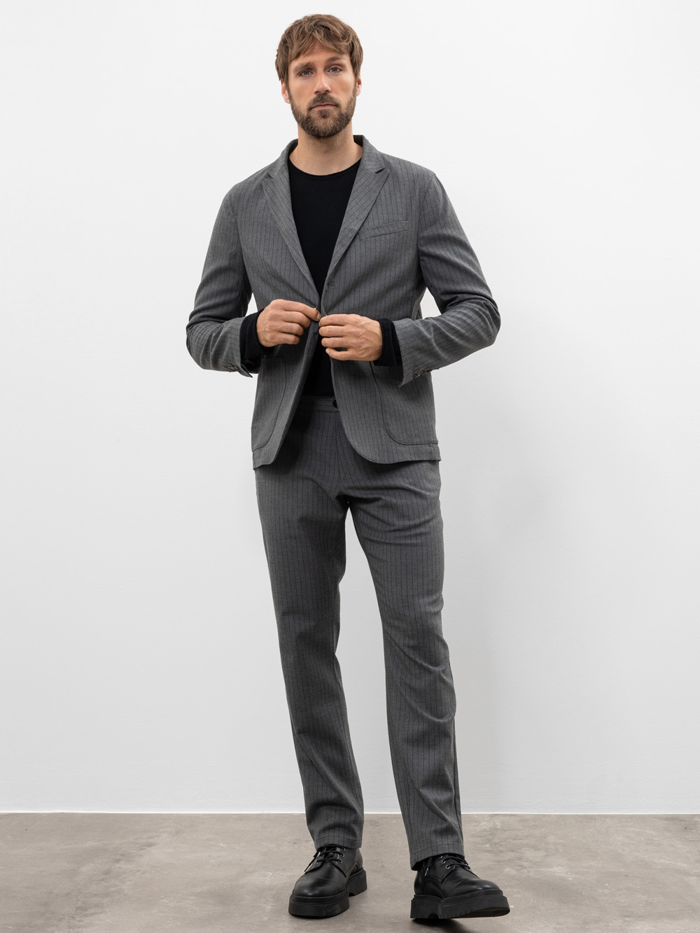 Ein Mann mit braunem Haar und Bart steht in einem Innenraum vor einer schlichten weißen Wand und trägt einen grauen Nadelstreifenanzug, ein schwarzes Hemd und schwarze Schuhe. Er knöpft seine Anzugsjacke zu und schaut in die Kamera.