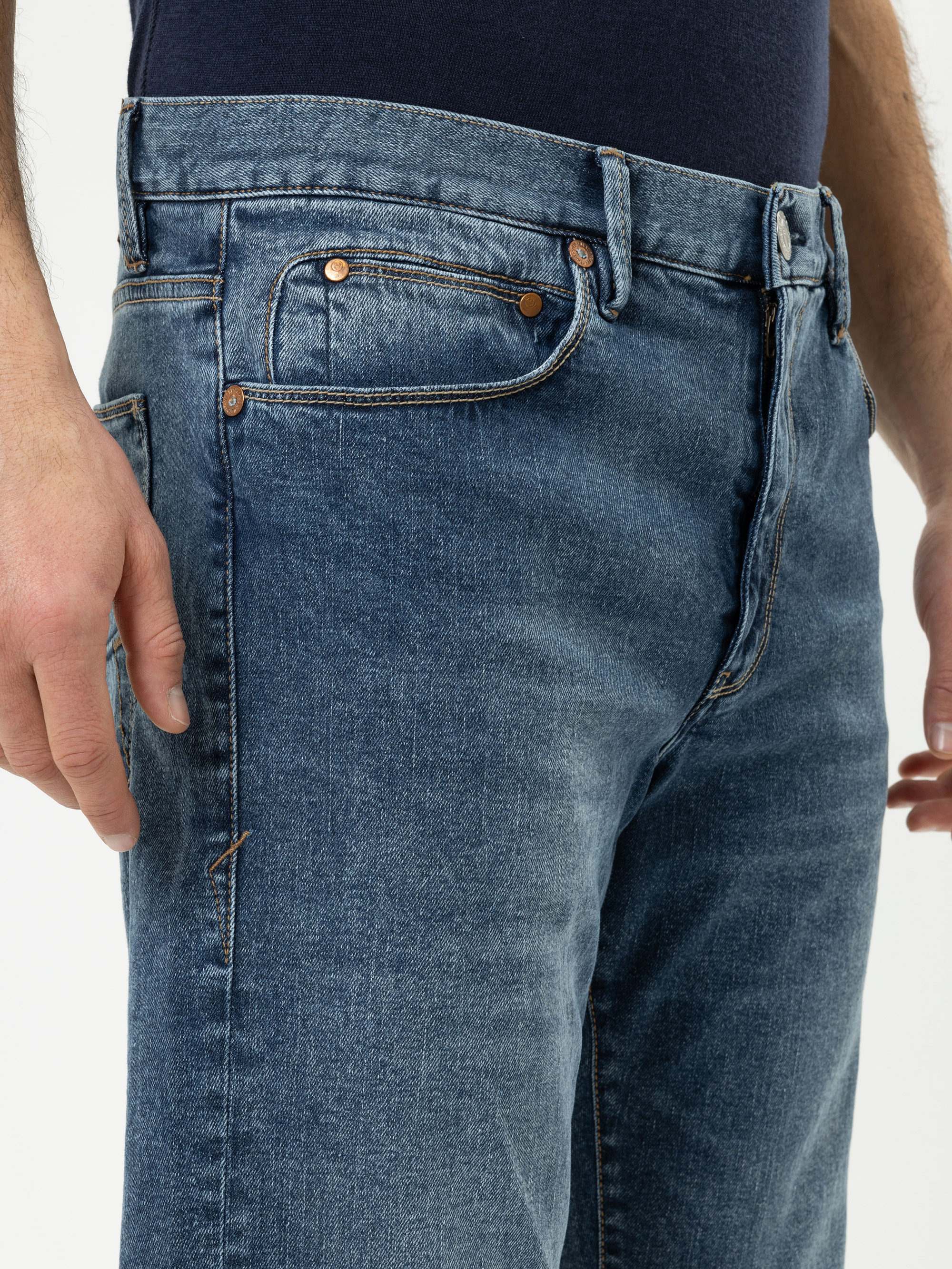Nahaufnahme einer Person, die eine blaue Jeans und ein dunkles Hemd trägt, wobei der Schwerpunkt auf der Taille und den Oberschenkeln liegt. Die Jeans hat kupferne Nieten und eine sichtbare Münztasche.