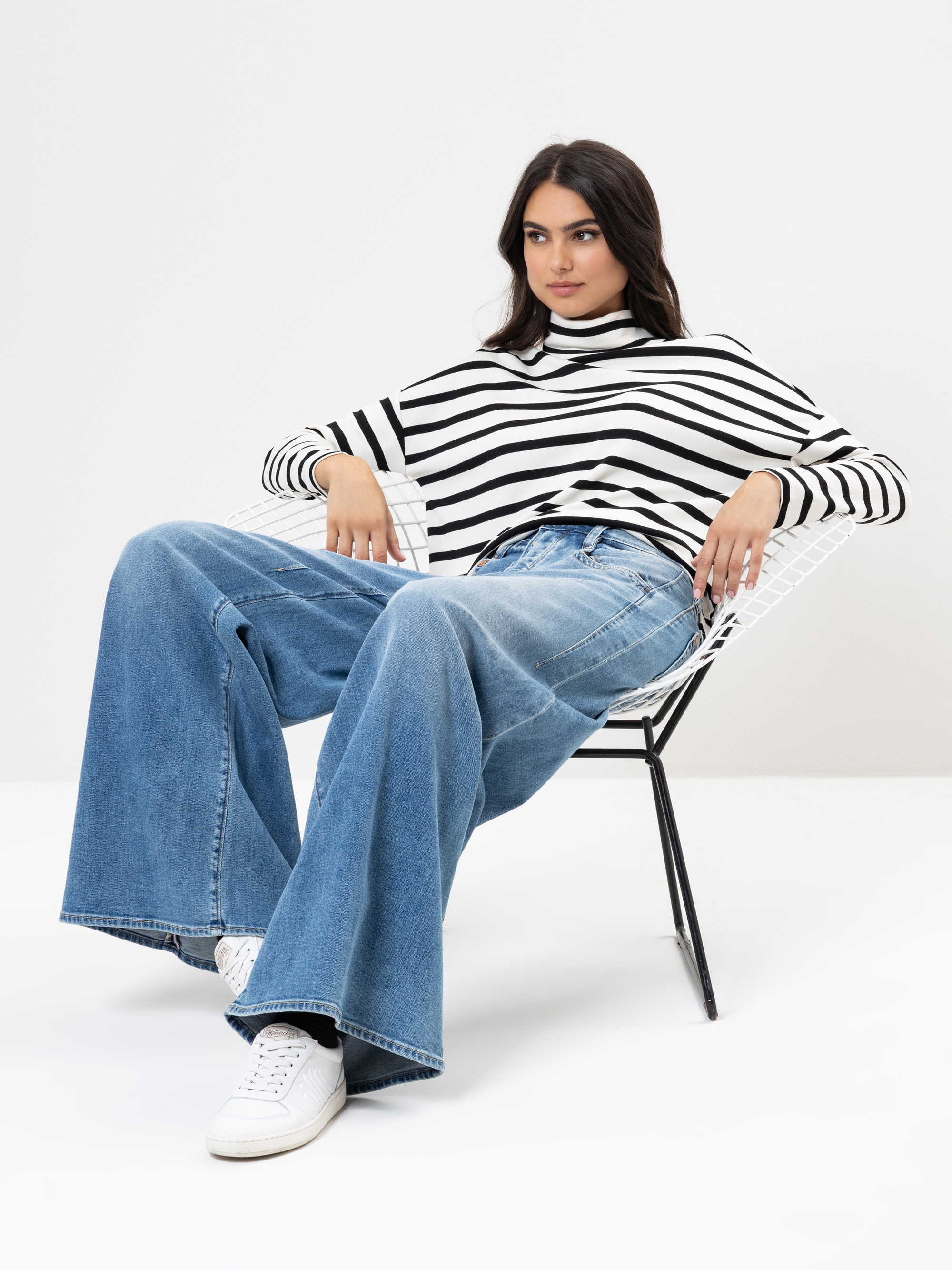 Eine Frau, die ein schwarz-weiß gestreiftes, langärmeliges Oberteil, eine weit geschnittene Bluejeans und weiße Turnschuhe trägt, sitzt entspannt auf einem modernen Drahtstuhl vor einem schlichten weißen Hintergrund.