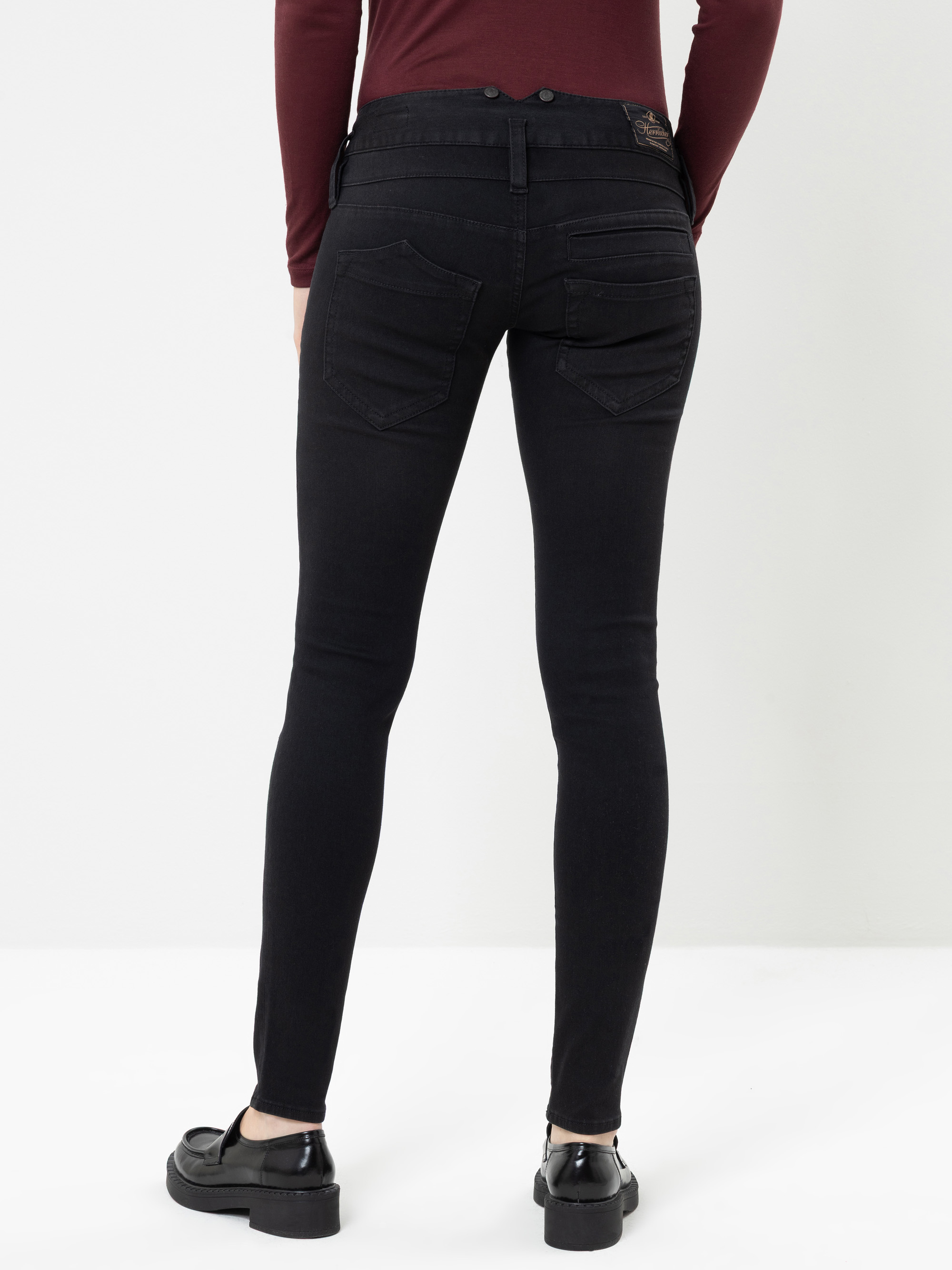 Eine Person, die eine schwarze Skinny-Jeans, ein burgunderrotes Langarm-Oberteil und schwarze Slipper trägt, steht mit dem Rücken zu einer weißen Wand.