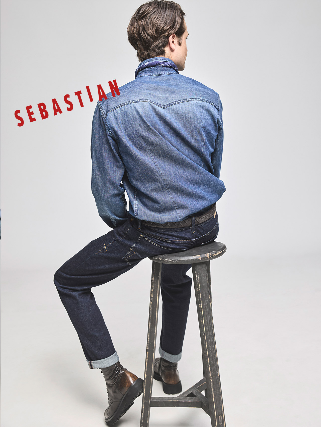 Ein Mann mit braunem Haar, der ein Jeanshemd, dunkle Jeans und Stiefel trägt, sitzt auf einem hohen Hocker und blickt zur Seite. Der Name SEBASTIAN steht in fetter roter Schrift auf der linken Seite des Bildes.