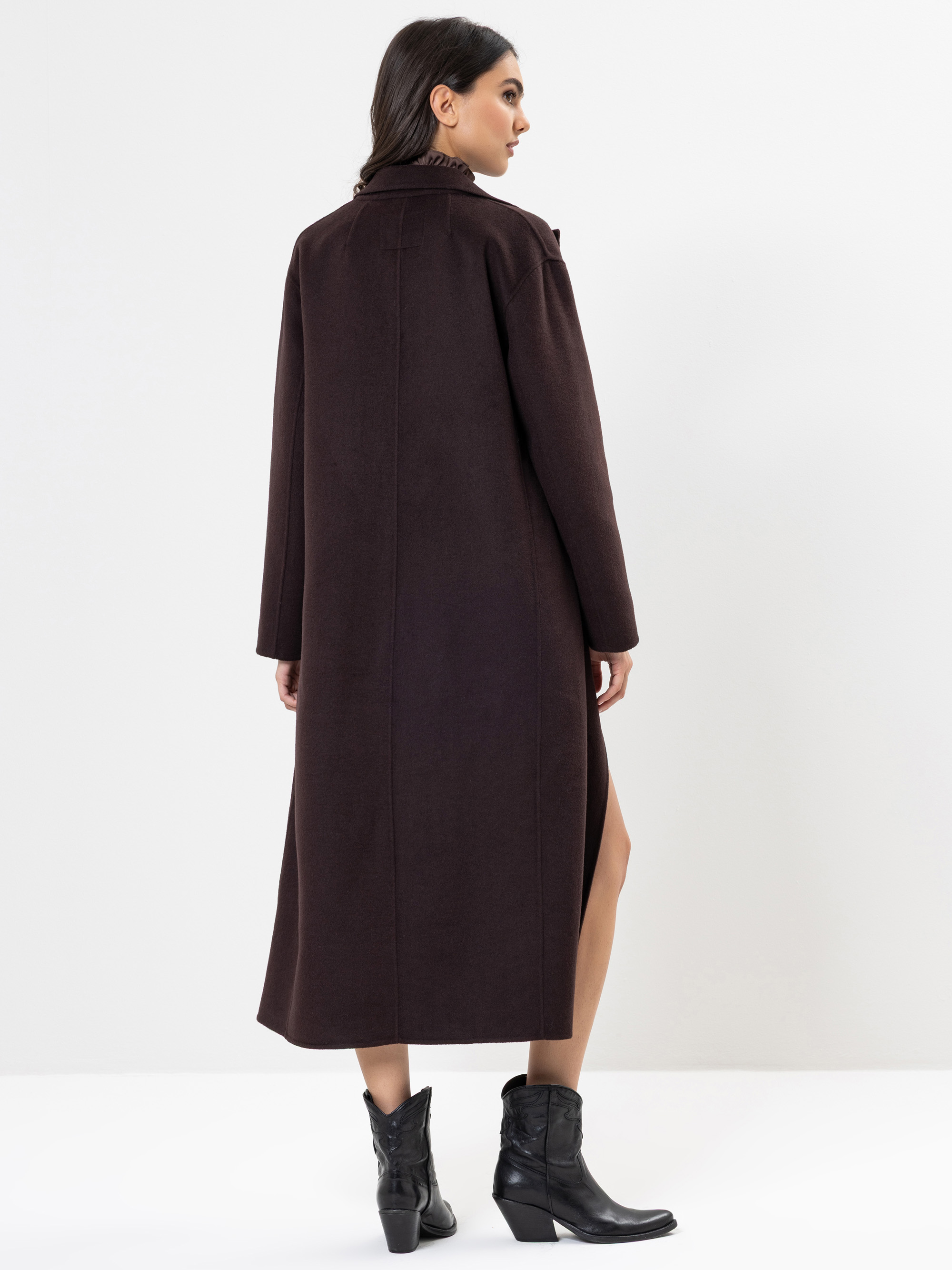 Frau, die mit dem Rücken zur Kamera steht und einen langen dunkelbraunen Mantel mit hohem Kragen und schwarzen Stiefeln trägt, vor einem schlichten weißen Hintergrund.