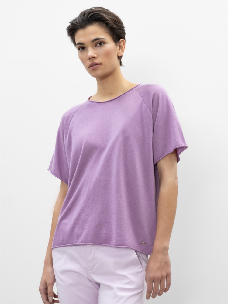 Eine Person mit kurzen dunklen Haaren, die ein locker sitzendes, kurzärmeliges lavendelfarbenes Oberteil und eine hellrosa Hose trägt, steht vor einem schlichten weißen Hintergrund.