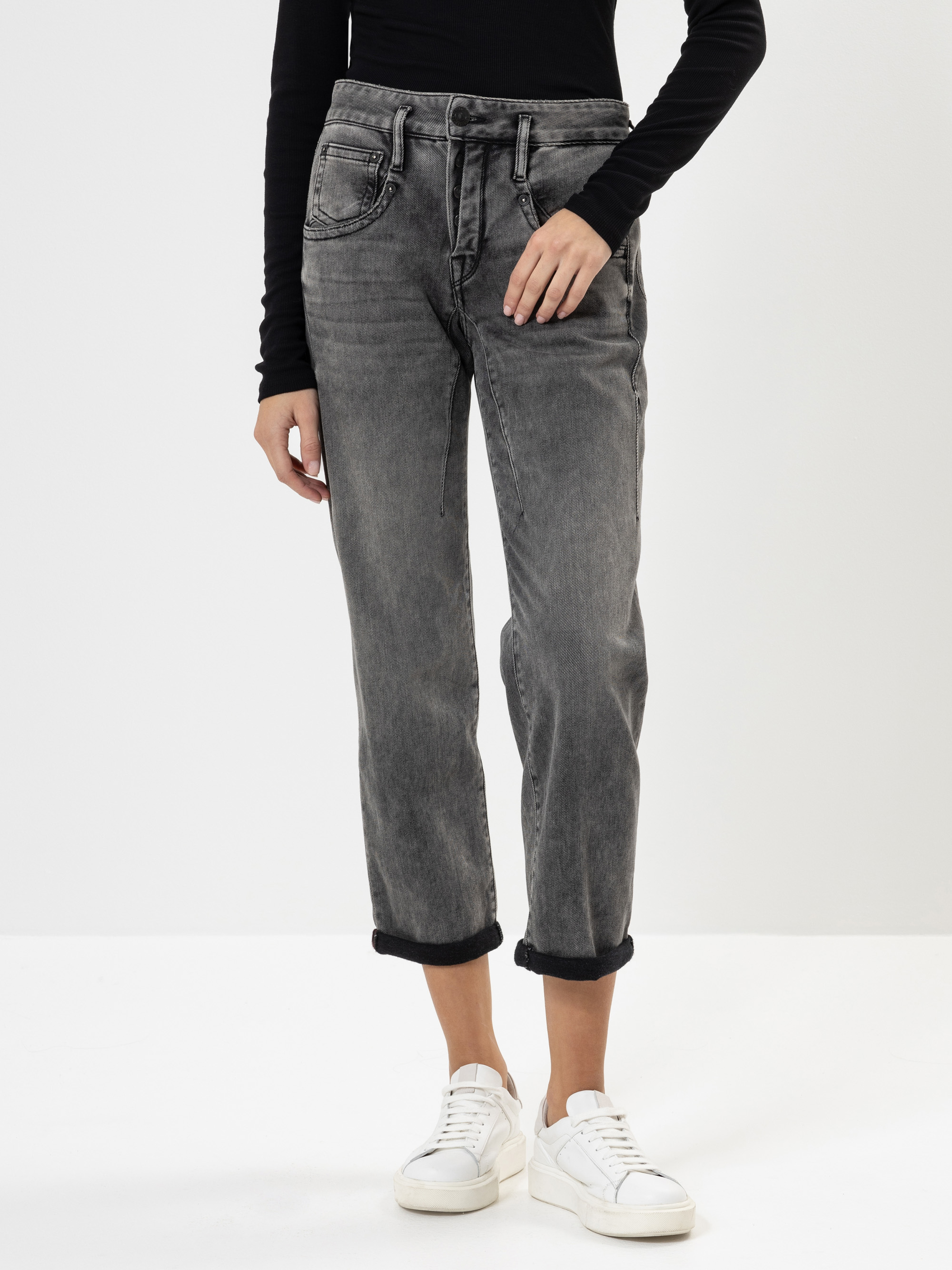 Person, die ein schwarzes langärmeliges Oberteil, graue Jeans mit Bündchen und weiße Turnschuhe trägt und vor einem schlichten weißen Hintergrund steht. Ihr Gesicht ist auf dem Bild nicht zu sehen.