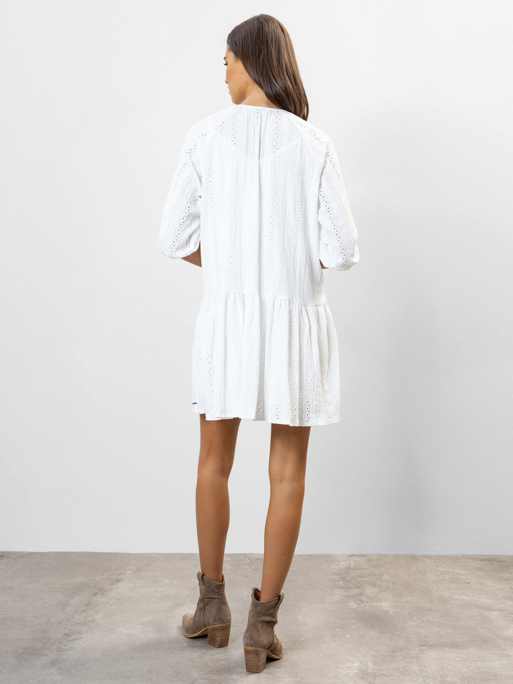 Eine Frau mit langen braunen Haaren steht mit dem Gesicht zur Wand. Sie trägt ein lockeres weißes Kleid mit Dreiviertelärmeln und braune Stiefeletten. Sie steht auf einem Betonboden vor einer schlichten weißen Wand.