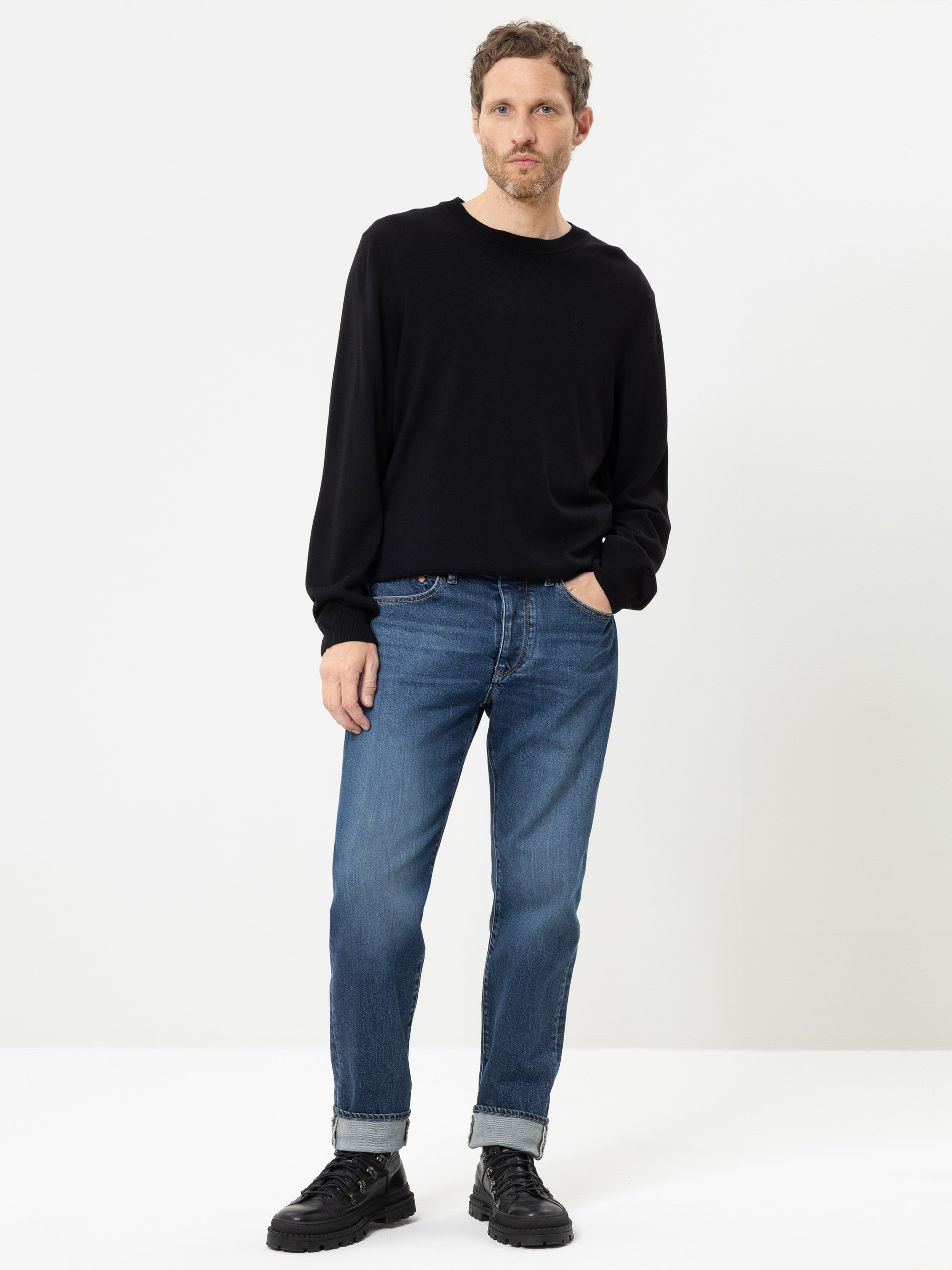 Vor einem weißen Hintergrund steht ein Mann, der ein schwarzes Langarmhemd, eine blaue Jeans mit Umschlag und schwarze Schuhe trägt; eine Hand hat er in der Tasche.