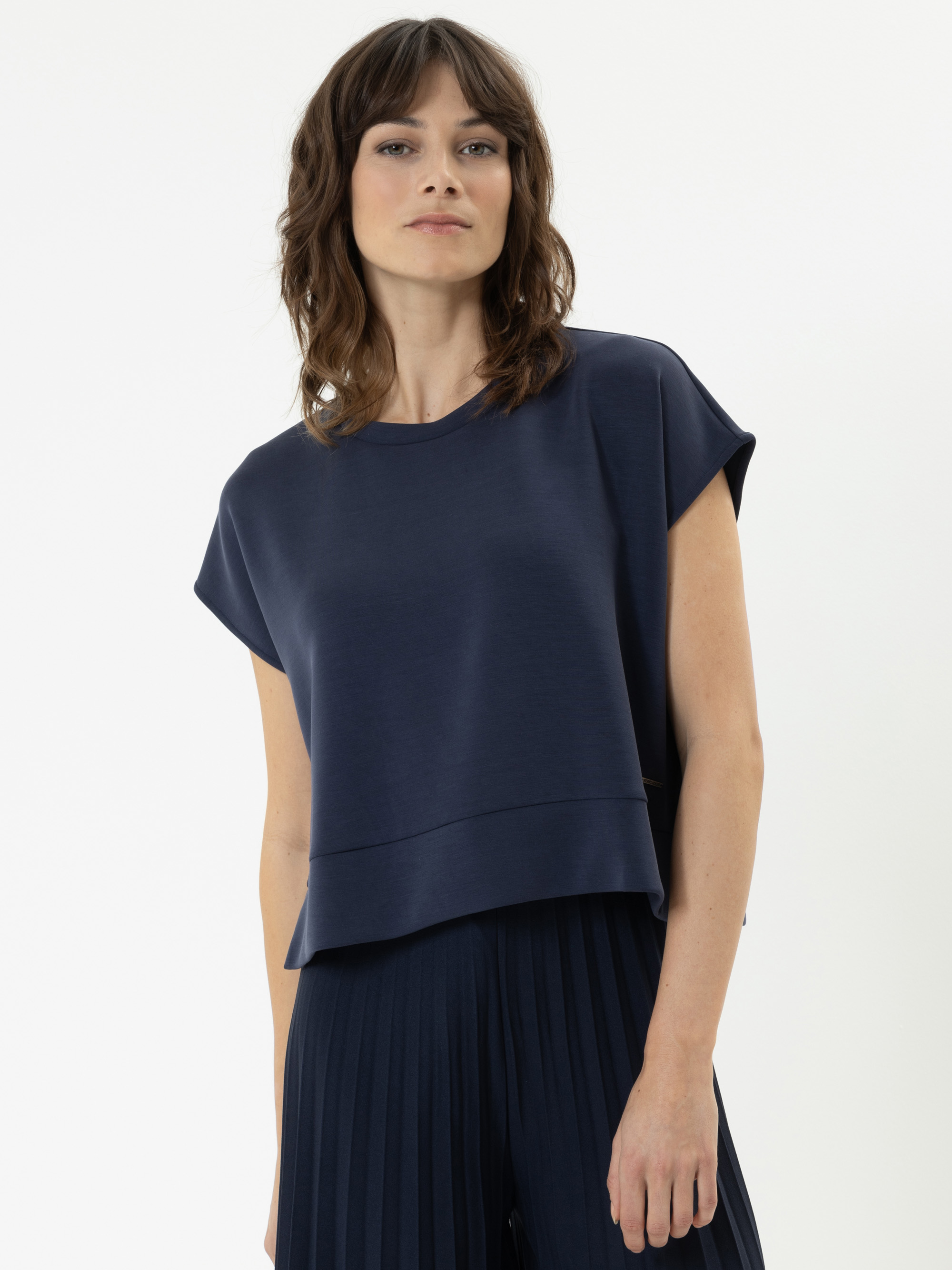 Eine Person mit gewelltem braunem Haar trägt ein marineblaues, kurzärmeliges, locker sitzendes Oberteil und eine dazu passende Bundfaltenhose vor einem schlichten weißen Hintergrund.