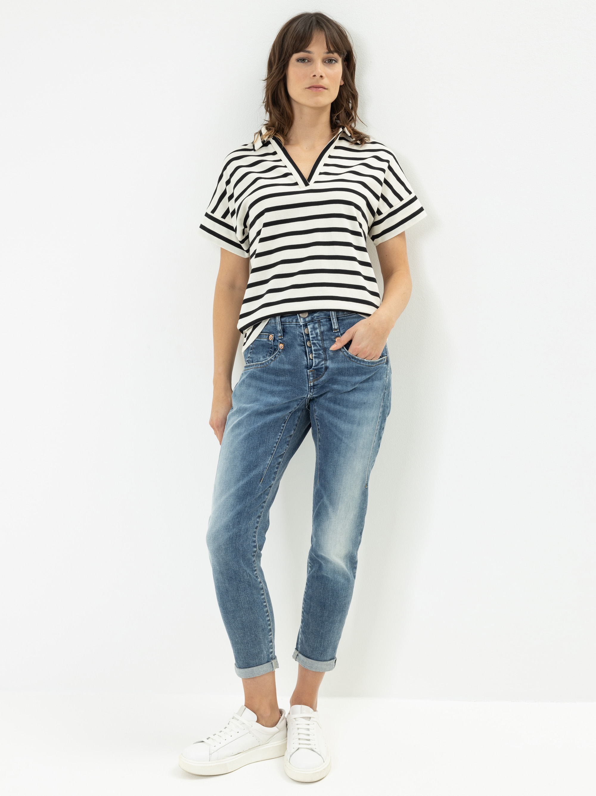 Eine Frau, die ein schwarz-weiß gestreiftes, kurzärmeliges Hemd, blaue Jeans mit Bündchen und weiße Turnschuhe trägt, steht vor einem schlichten weißen Hintergrund und hat eine Hand in der Tasche.