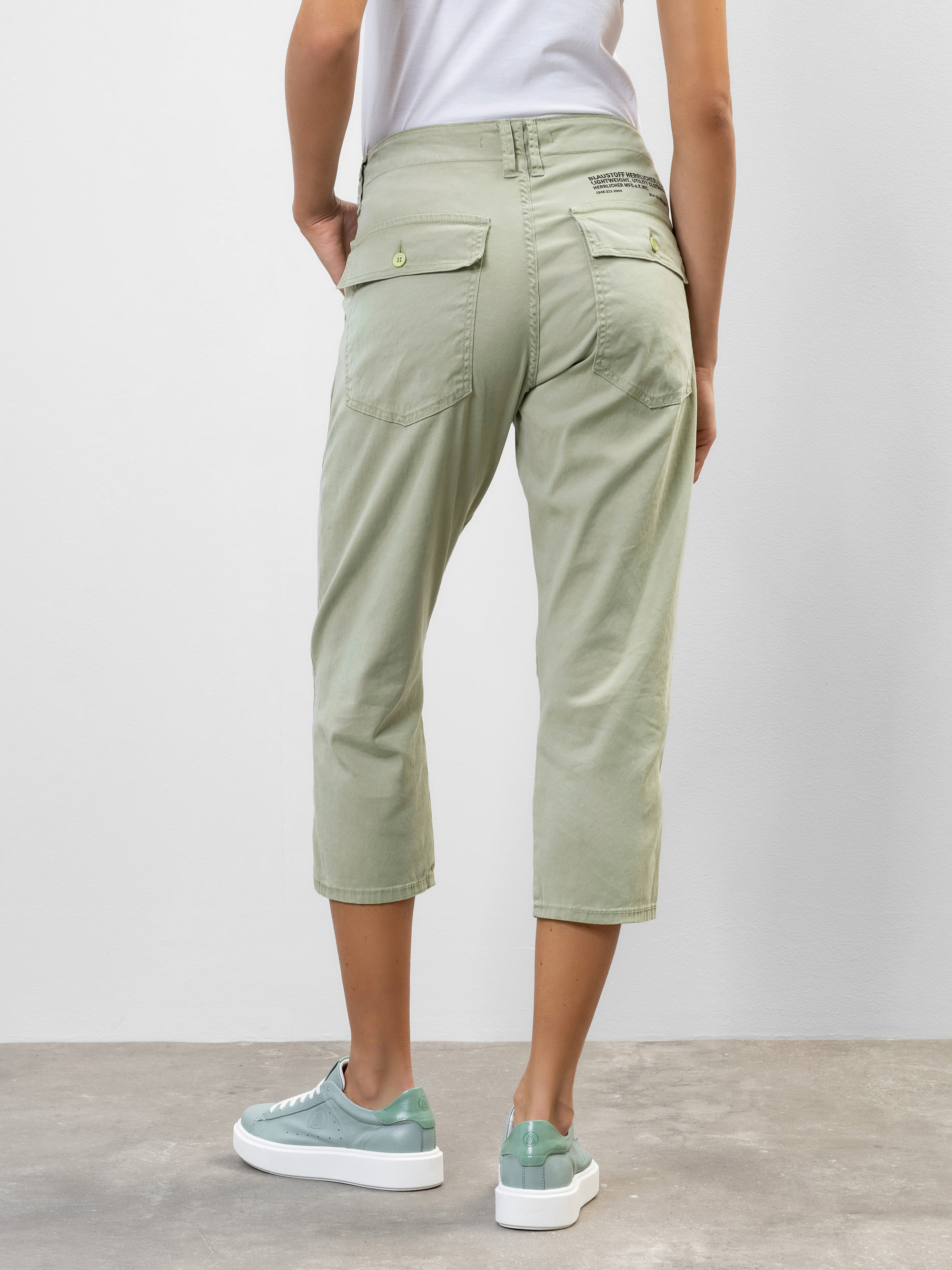 Eine Person, die in einem Haus steht, von hinten gesehen, trägt eine hellgrüne, kurze Hose und grüne Turnschuhe. Die Hose hat zwei Gesäßtaschen mit Knöpfen. Die Umgebung besteht aus einer schlichten hellen Wand und einem nackten Boden.