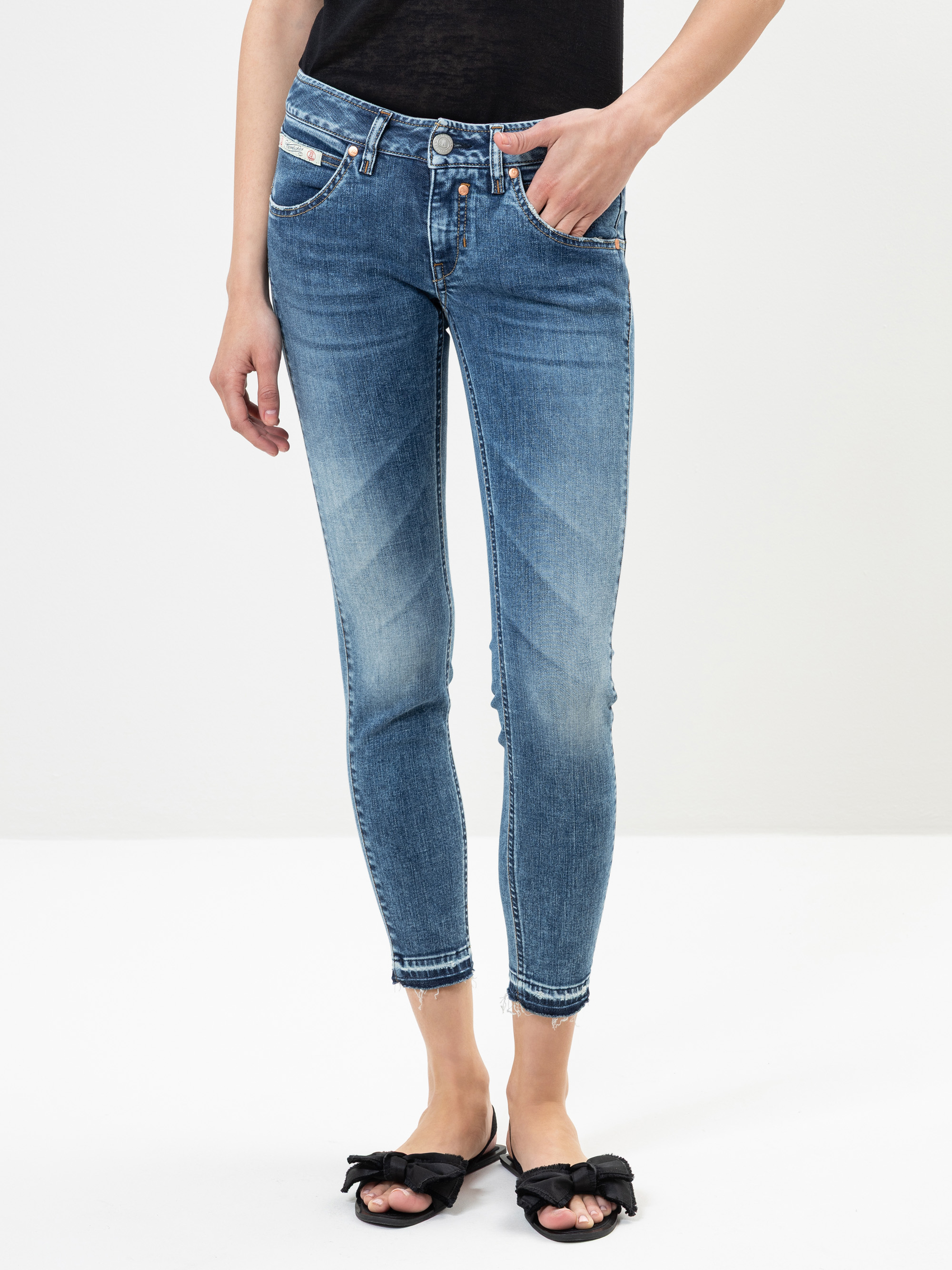 Eine Person, die ein schwarzes Oberteil, blaue Röhrenjeans mit verblassten Details und schwarze Gleitsandalen mit Schleifen trägt, steht vor einem schlichten weißen Hintergrund.