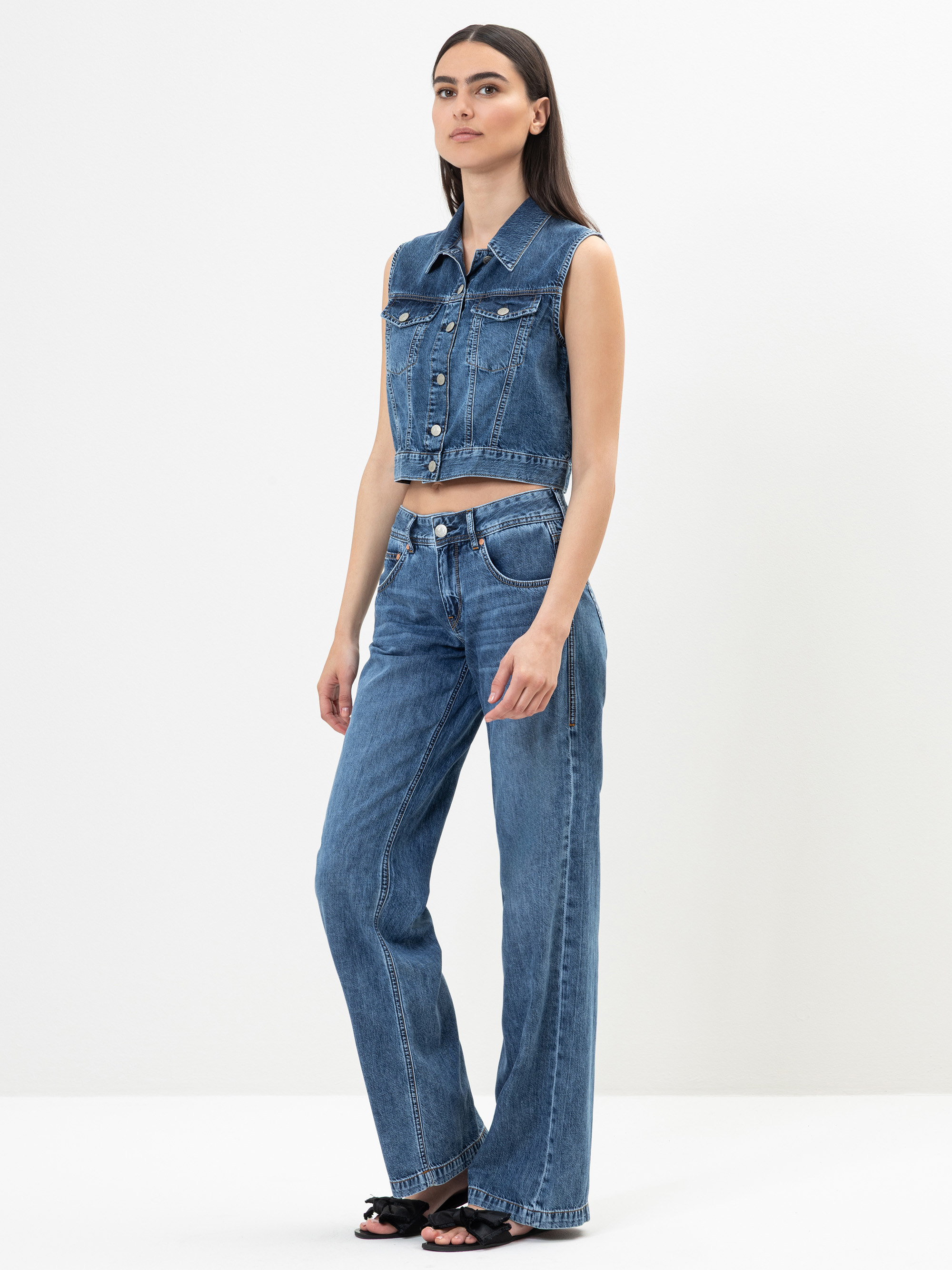 Eine Frau steht in einem Studio und trägt eine ärmellose blaue Jeansweste und eine dazu passende Jeans mit hoher Taille und weitem Bein, die an der Taille ausgeschnitten ist. Sie hat langes dunkles Haar und trägt schwarze, offene Absätze.