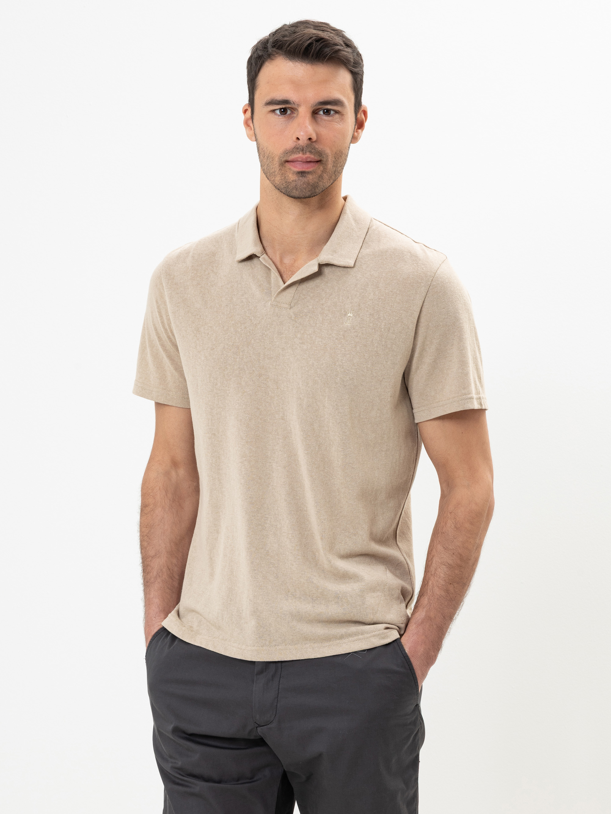 Ein Mann mit kurzen dunklen Haaren und gestutztem Bart trägt ein beiges Polohemd und eine dunkle Hose. Er steht vor einem schlichten weißen Hintergrund, hat die Hände in den Taschen und blickt mit neutraler Miene in die Kamera.