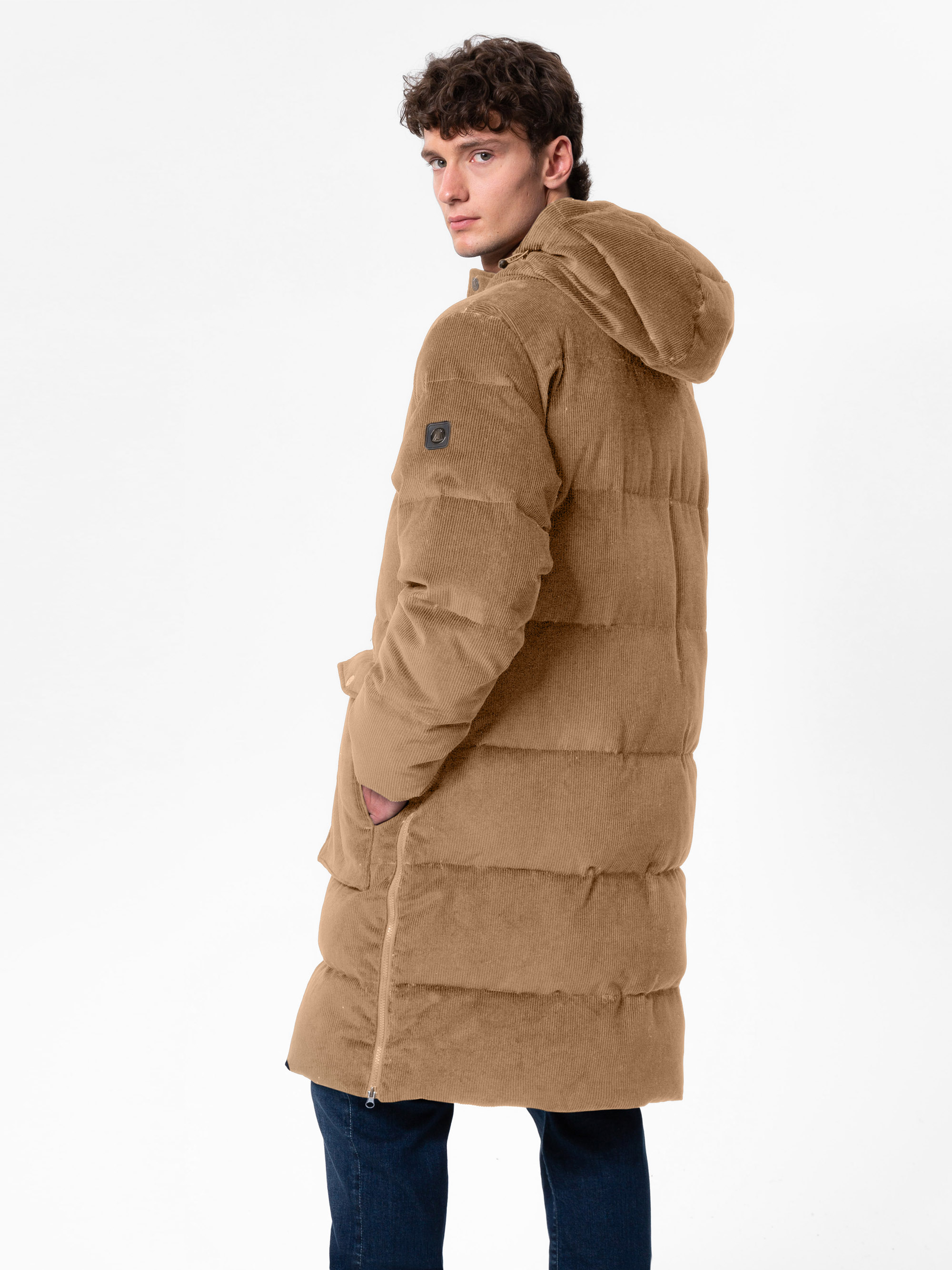 Ein junger Mann mit lockigem Haar, der einen langen, hellbraunen Mantel mit Kapuze trägt und über seine Schulter schaut. Er hat eine Hand in der Tasche und steht vor einem schlichten weißen Hintergrund.