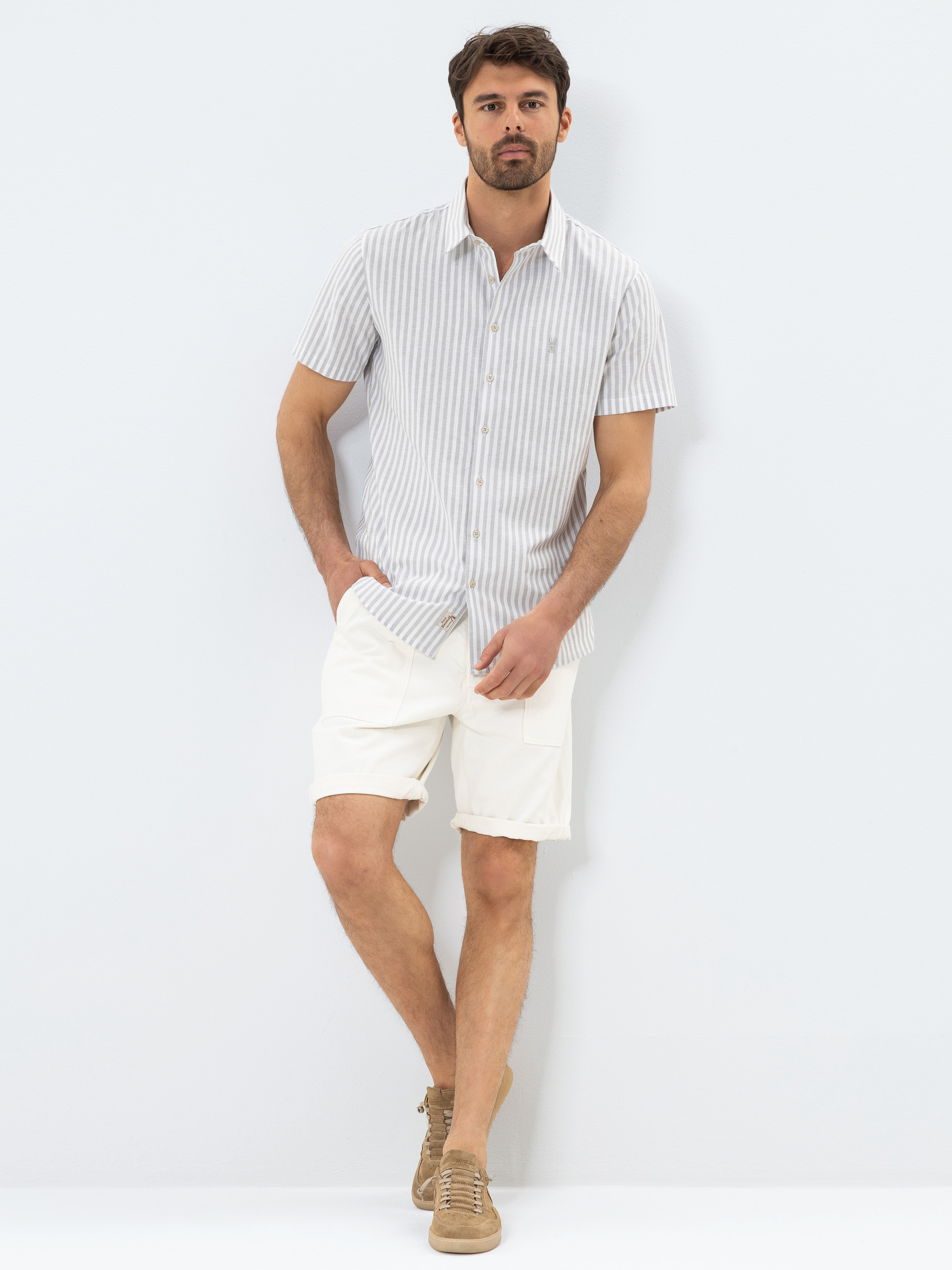 Ein Mann steht vor einem weißen Hintergrund und trägt ein kurzärmeliges, hellgrau-weiß gestreiftes Button-up-Hemd, weiße Shorts und hellbraune Turnschuhe. Eine Hand steckt in seiner Hosentasche und er blickt direkt in die Kamera.