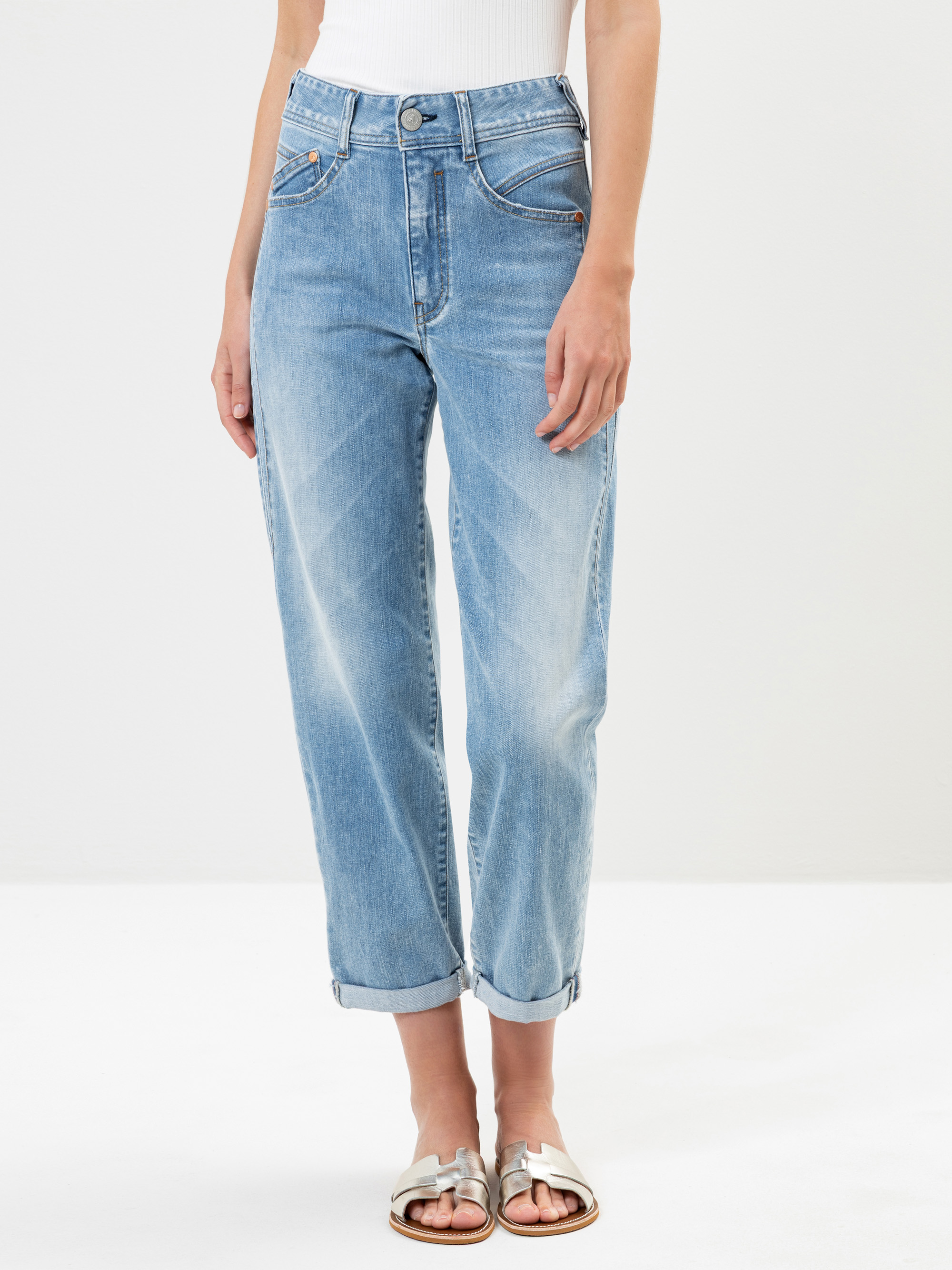 Eine Person in hellen Jeans mit Bündchen und einem weißen ärmellosen Oberteil steht vor einem schlichten Hintergrund. Sie trägt außerdem weiße Sandalen. Ihr Gesicht ist auf dem Bild nicht zu sehen.