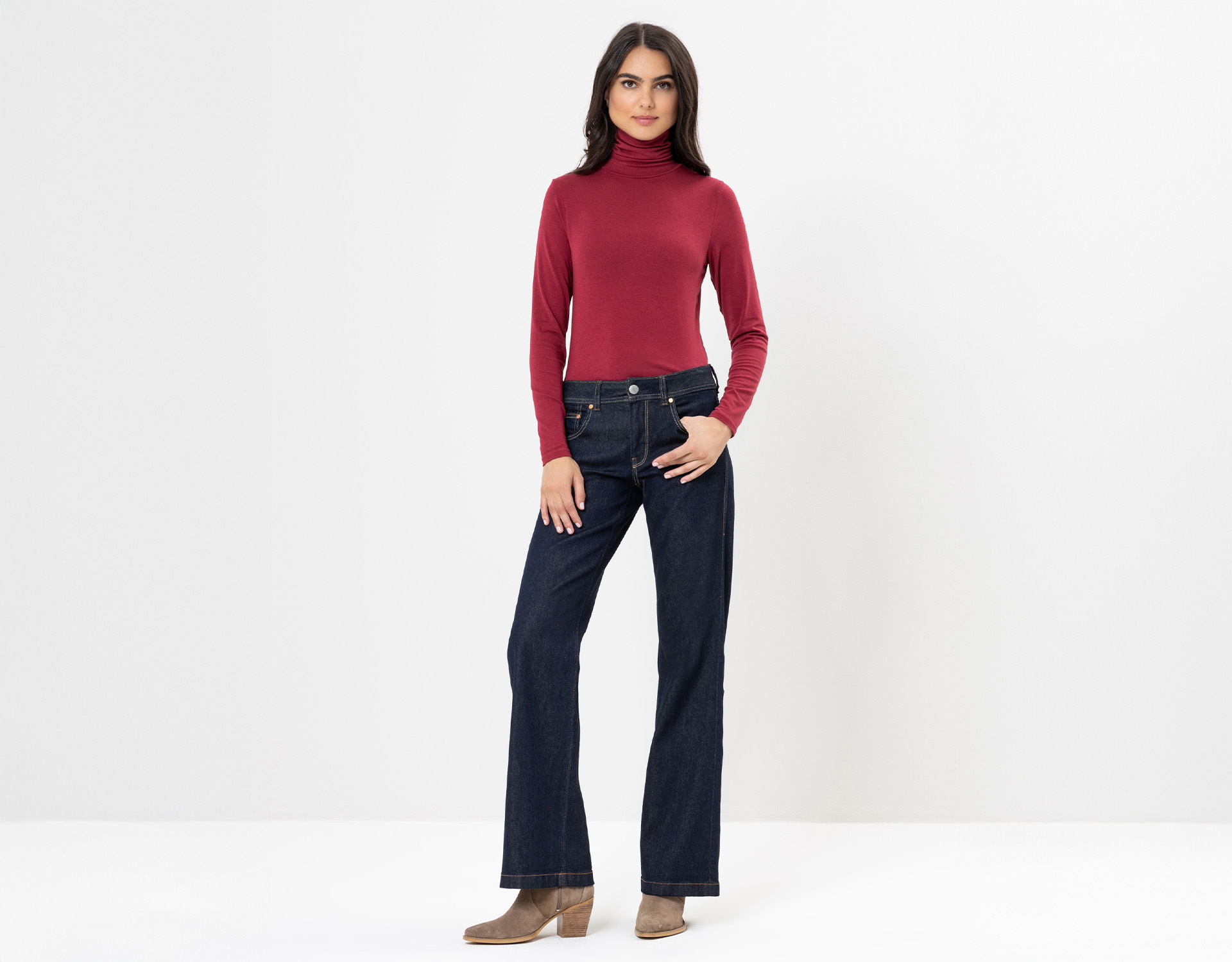 Eine Frau steht vor einem weißen Hintergrund und trägt einen roten Rollkragenpullover, dunkelblaue Jeans und hellbraune Stiefeletten. Sie blickt nach vorne, eine Hand in der Tasche, die andere an der Seite liegend.