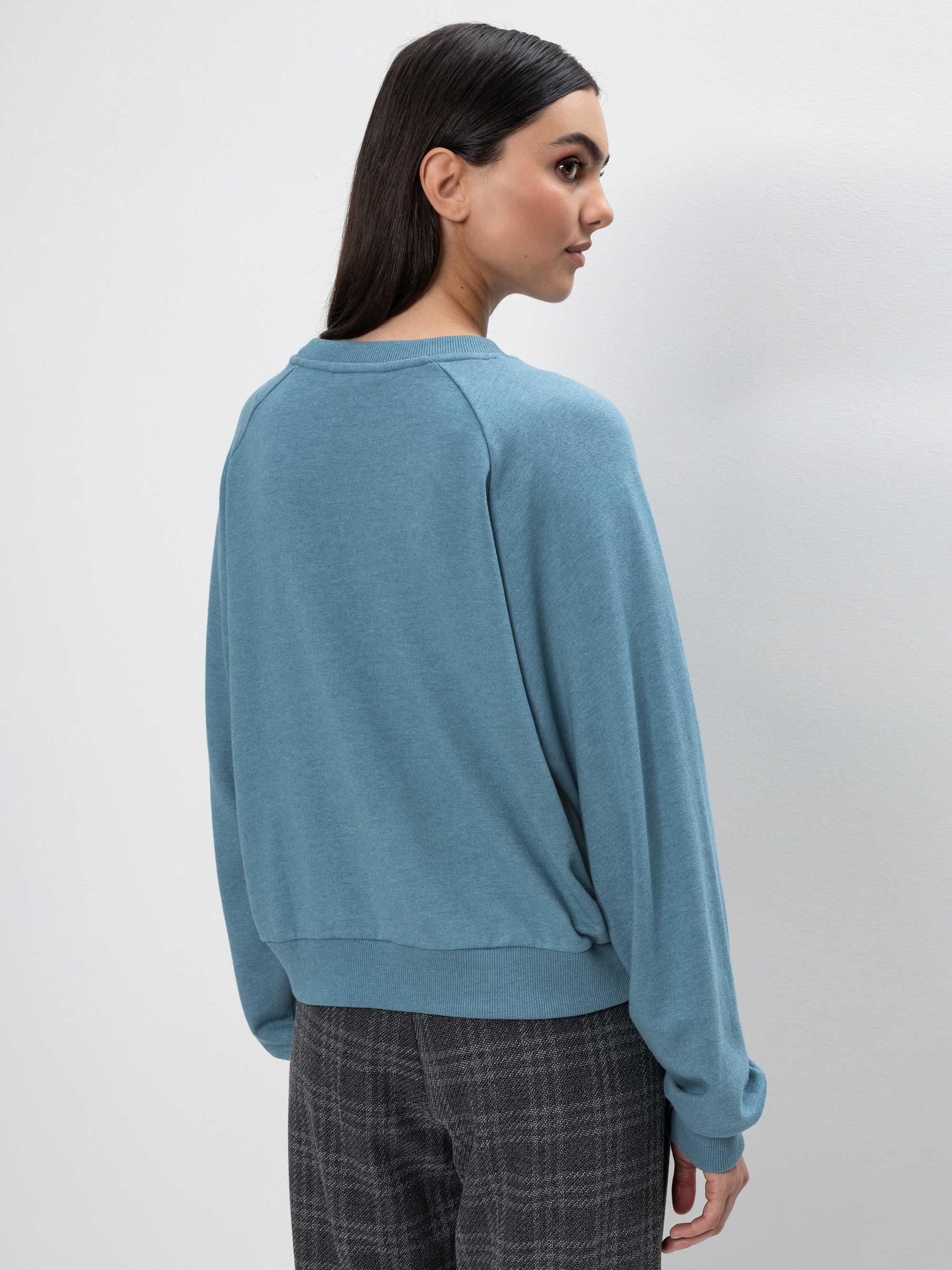 Eine Frau mit langen dunklen Haaren trägt ein hellblaues Sweatshirt und eine grau karierte Hose. Sie steht vor einem schlichten hellen Hintergrund, schaut nach links und wendet sich ab.