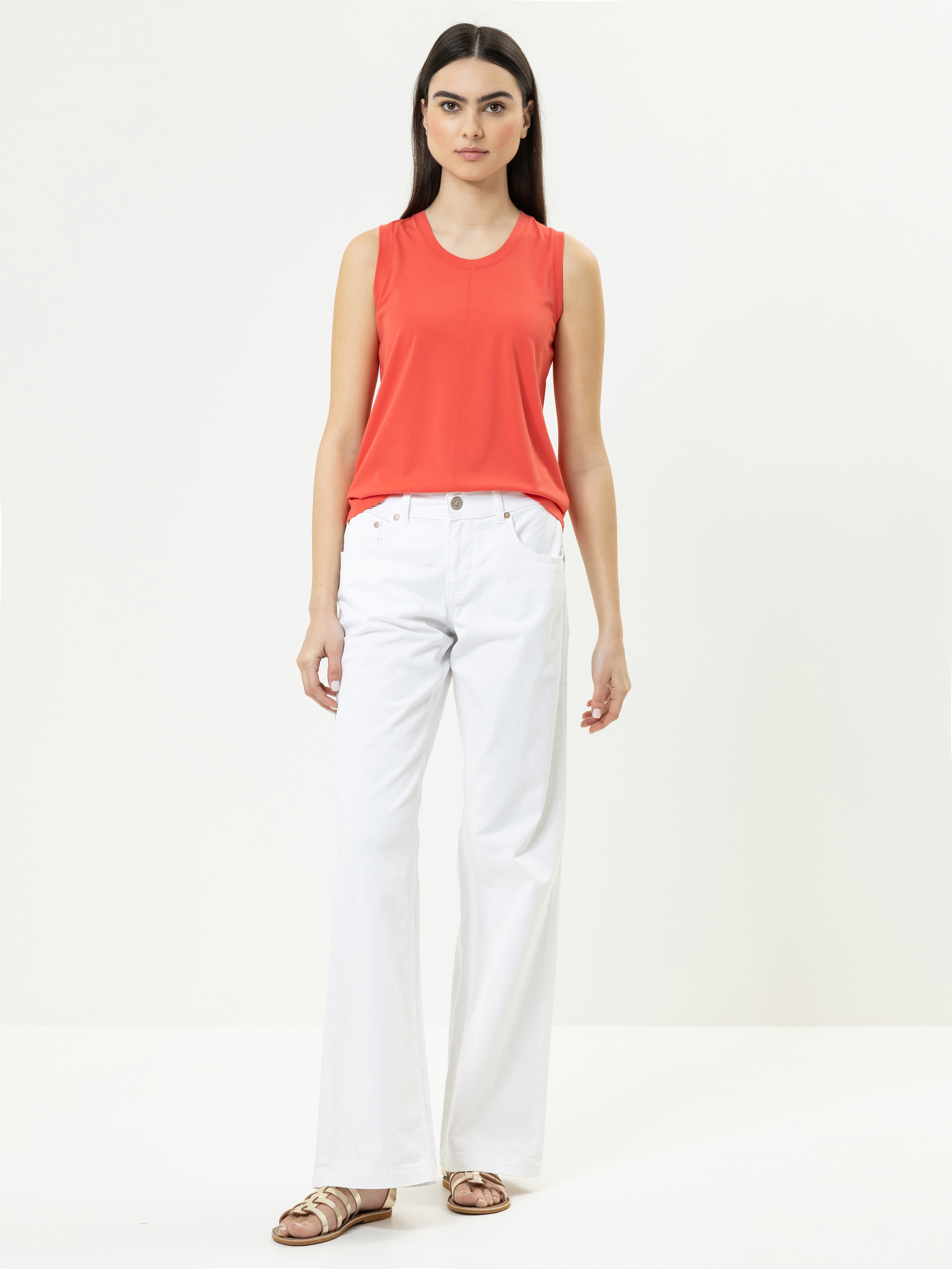 Eine Frau steht vor einem schlichten weißen Hintergrund und trägt ein ärmelloses korallenrotes Oberteil, weiße Jeans mit hoher Taille und goldene Sandalen. Sie hat langes dunkles Haar und einen neutralen Gesichtsausdruck.