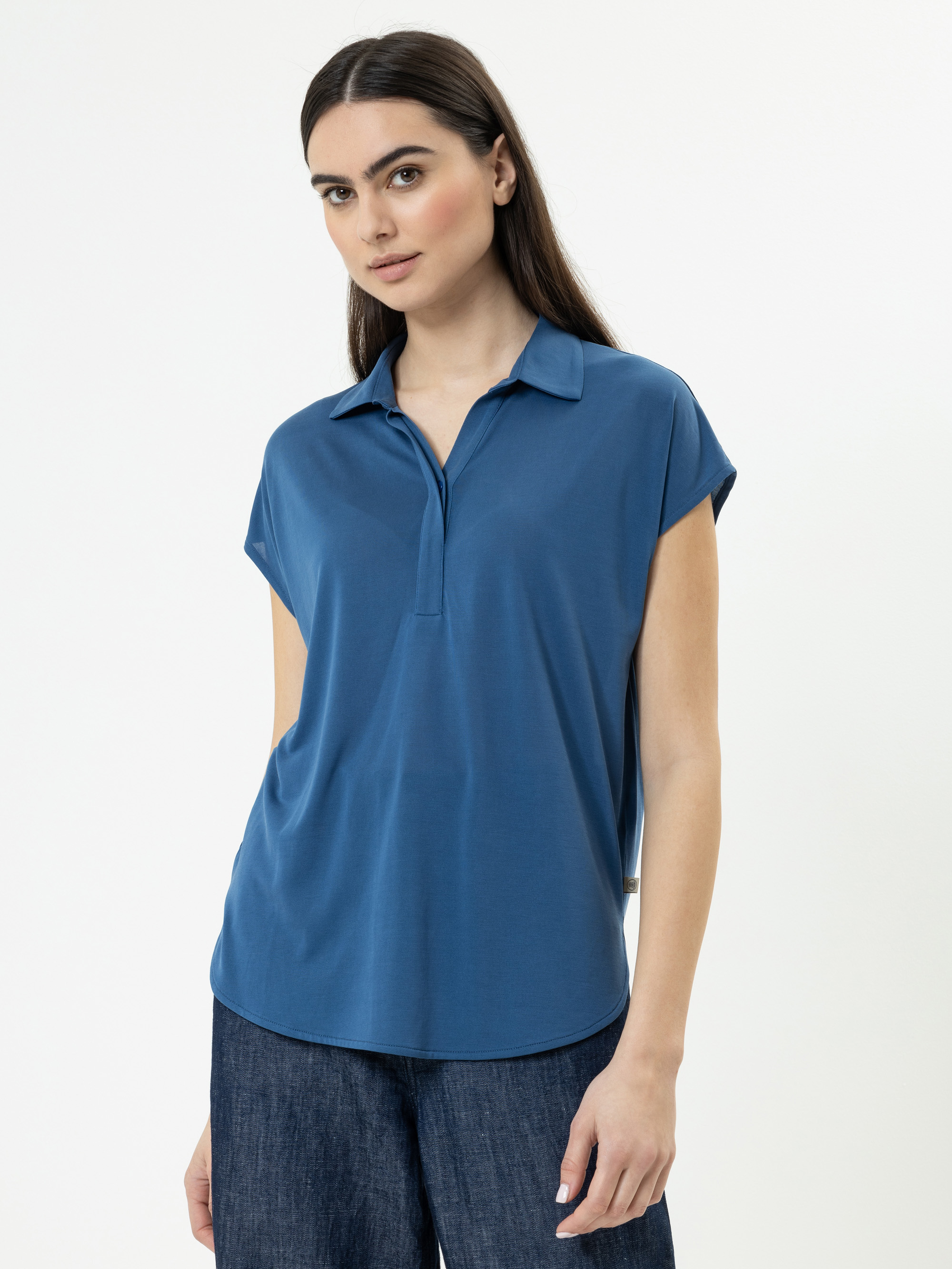 Eine Frau mit langen dunklen Haaren trägt ein kurzärmeliges, blaues Poloshirt mit Kragen und dunkle Jeans und steht vor einem schlichten weißen Hintergrund. Sie blickt leicht zur Seite und hat einen entspannten Gesichtsausdruck.